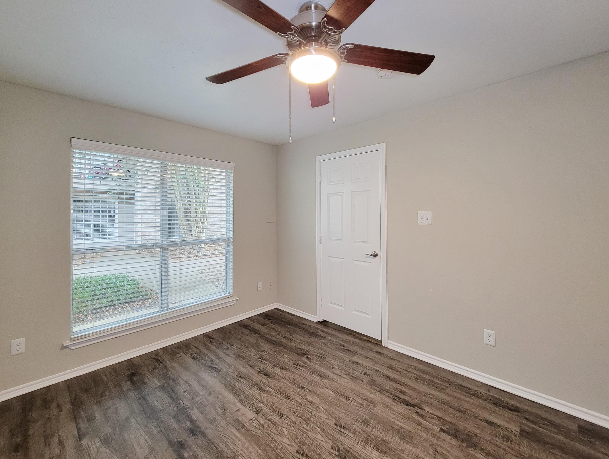 Empty room with light gray walls and medium brown wooden flooring. A ceiling fan with light is mounted in the center. The room features a large window with white blinds, allowing natural light to enter. A white door leads to another space, and there are no furnishings present.