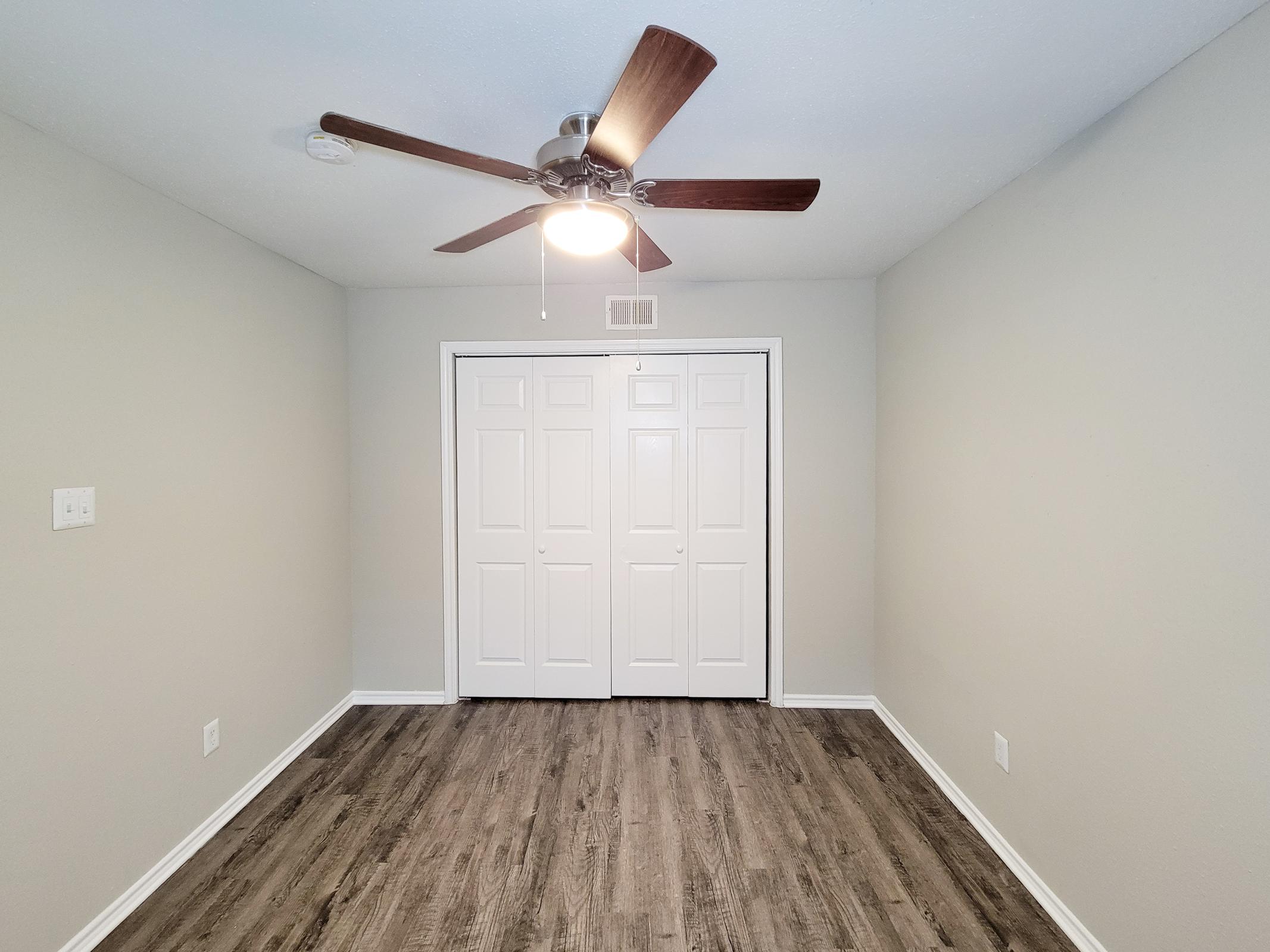 A spacious, neutral-colored room with a ceiling fan and wood-like flooring. The walls are light gray, and there is a closed double closet with white doors. The room is well-lit and organized, suitable for various uses such as a bedroom or office.