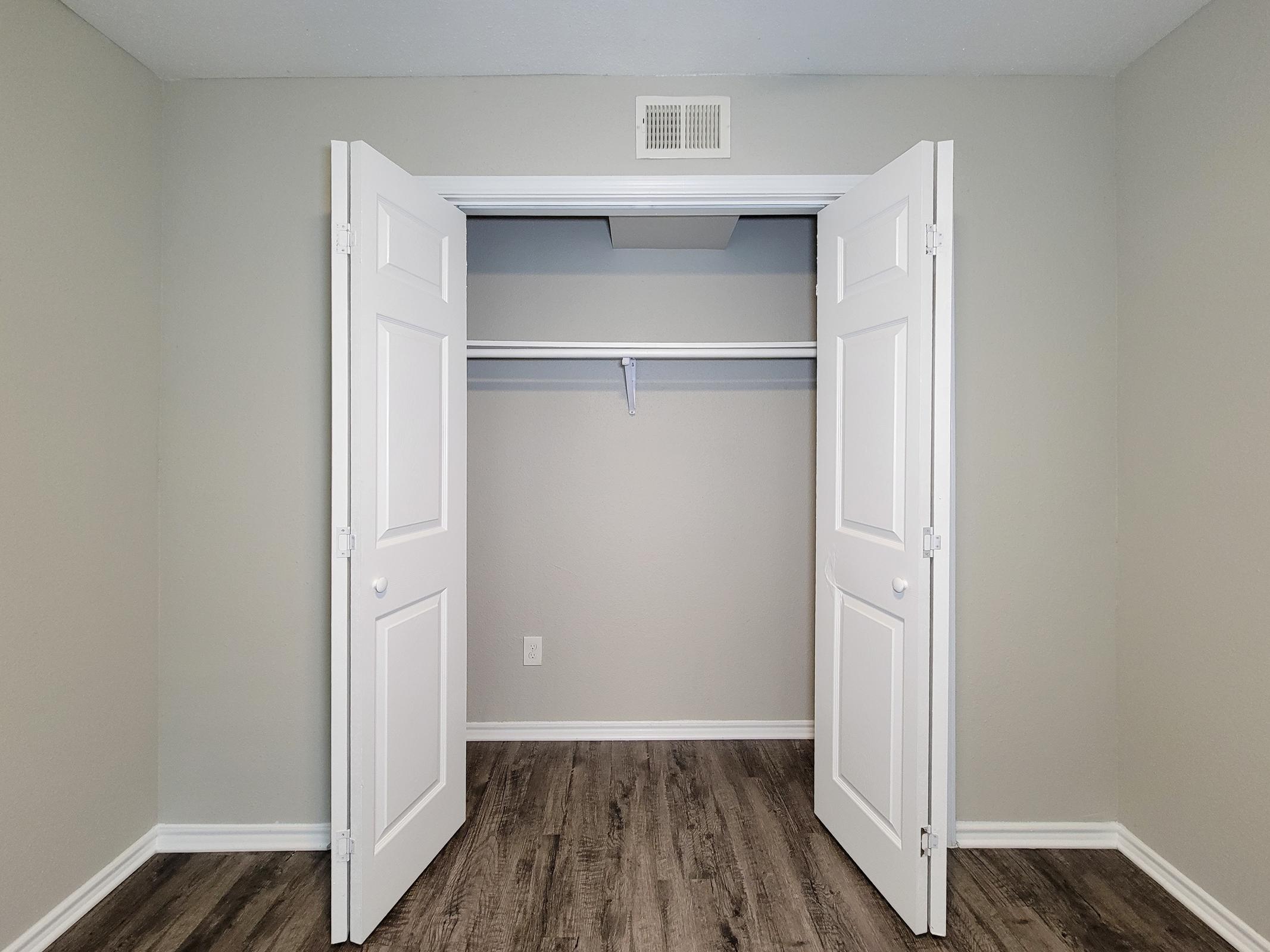 A spacious, empty closet with double white doors open, revealing a simple shelving system. The walls are painted a light gray, and the floor features a dark wood-like laminate. The room has neutral lighting and minimalist decor.