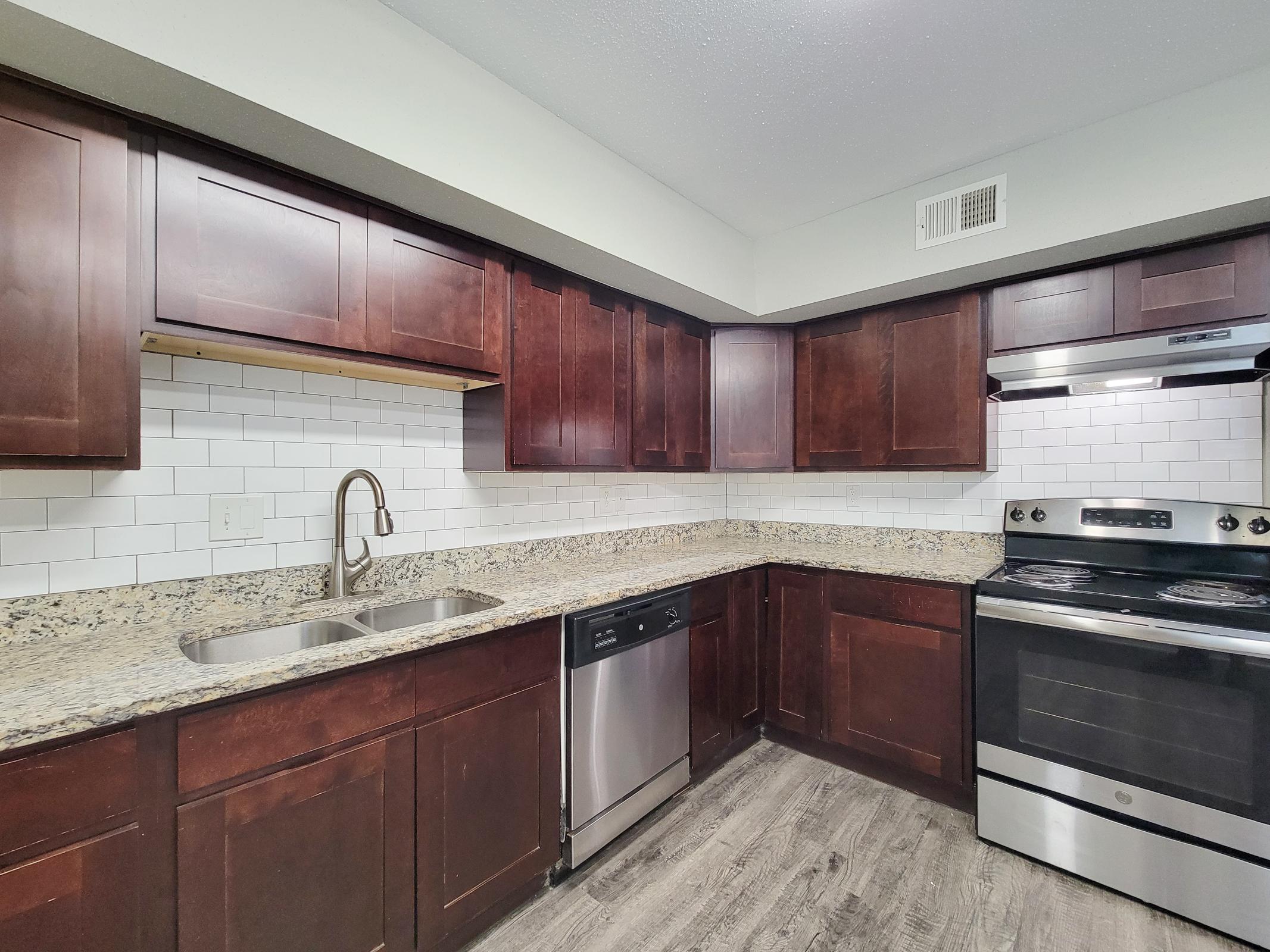 Modern kitchen featuring dark wood cabinetry, granite countertops, stainless steel appliances, and a white tile backsplash. The space includes a double sink, a dishwasher, and an oven. Natural light enhances the inviting atmosphere of the well-designed layout.