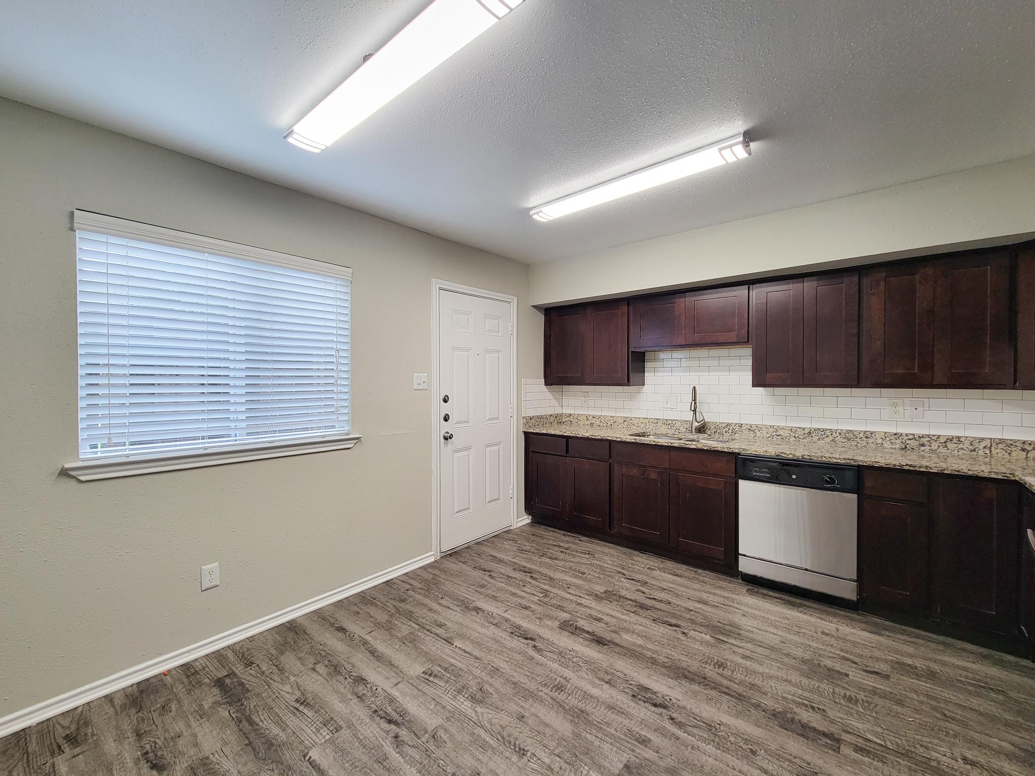 A modern kitchen featuring dark wooden cabinets, a light-colored granite countertop, and stainless steel appliances. There is a window with white blinds allowing natural light in, and the floor is covered with laminate wood planks. The entrance door is visible on the right.