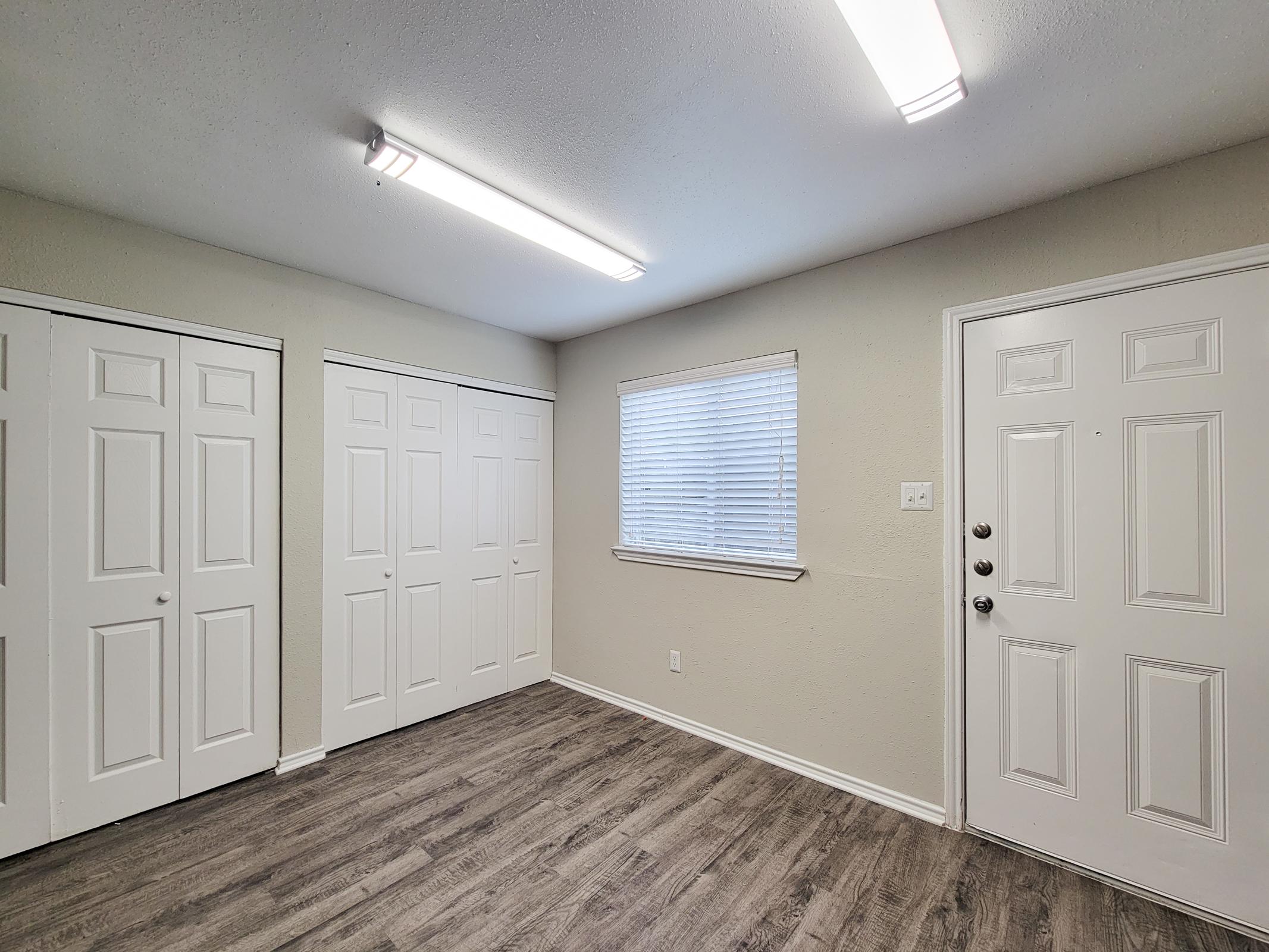 Interior view of a room featuring light-colored walls, a window with blinds, a door with a handle, and two sets of white double doors. The flooring is a light wood laminate, and ceiling lights provide ample illumination. The space appears clean and well-maintained, suitable for various uses.