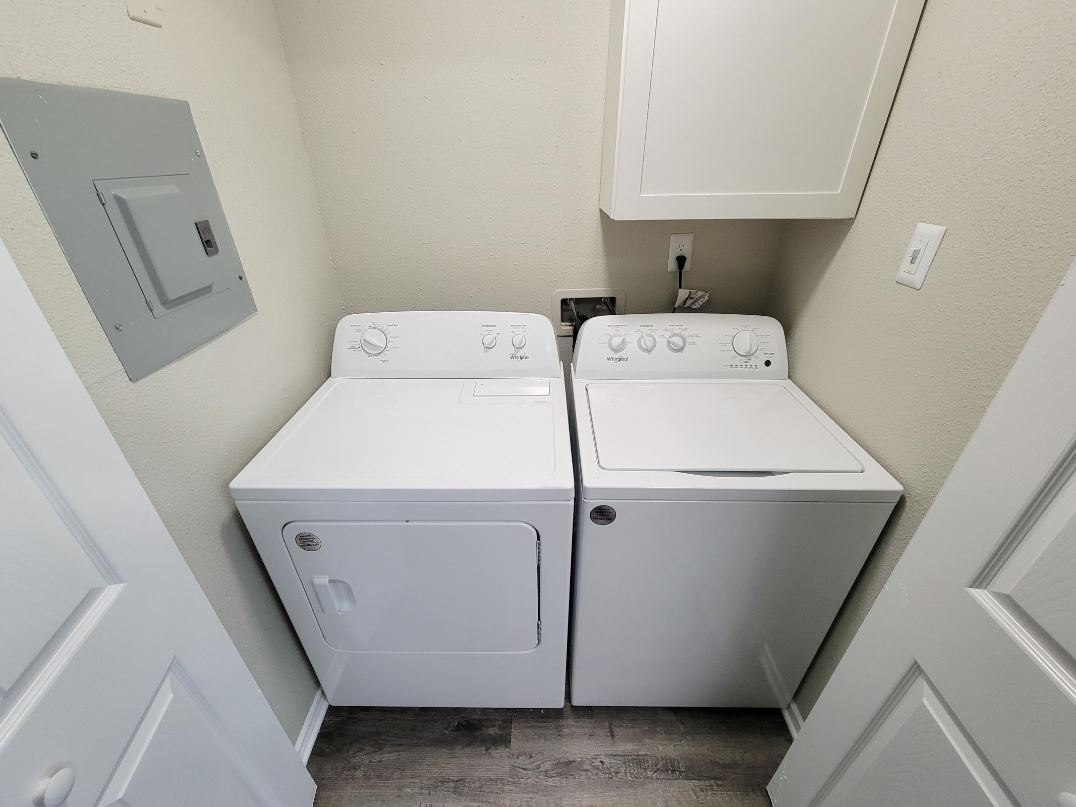 A laundry room featuring a white washing machine and a dryer next to each other. The appliances have control dials on the front. There's a light-colored wall behind them, and a white cabinet above the washer. A gray electrical panel is mounted on the wall to the left. The floor is a wood-like laminate.