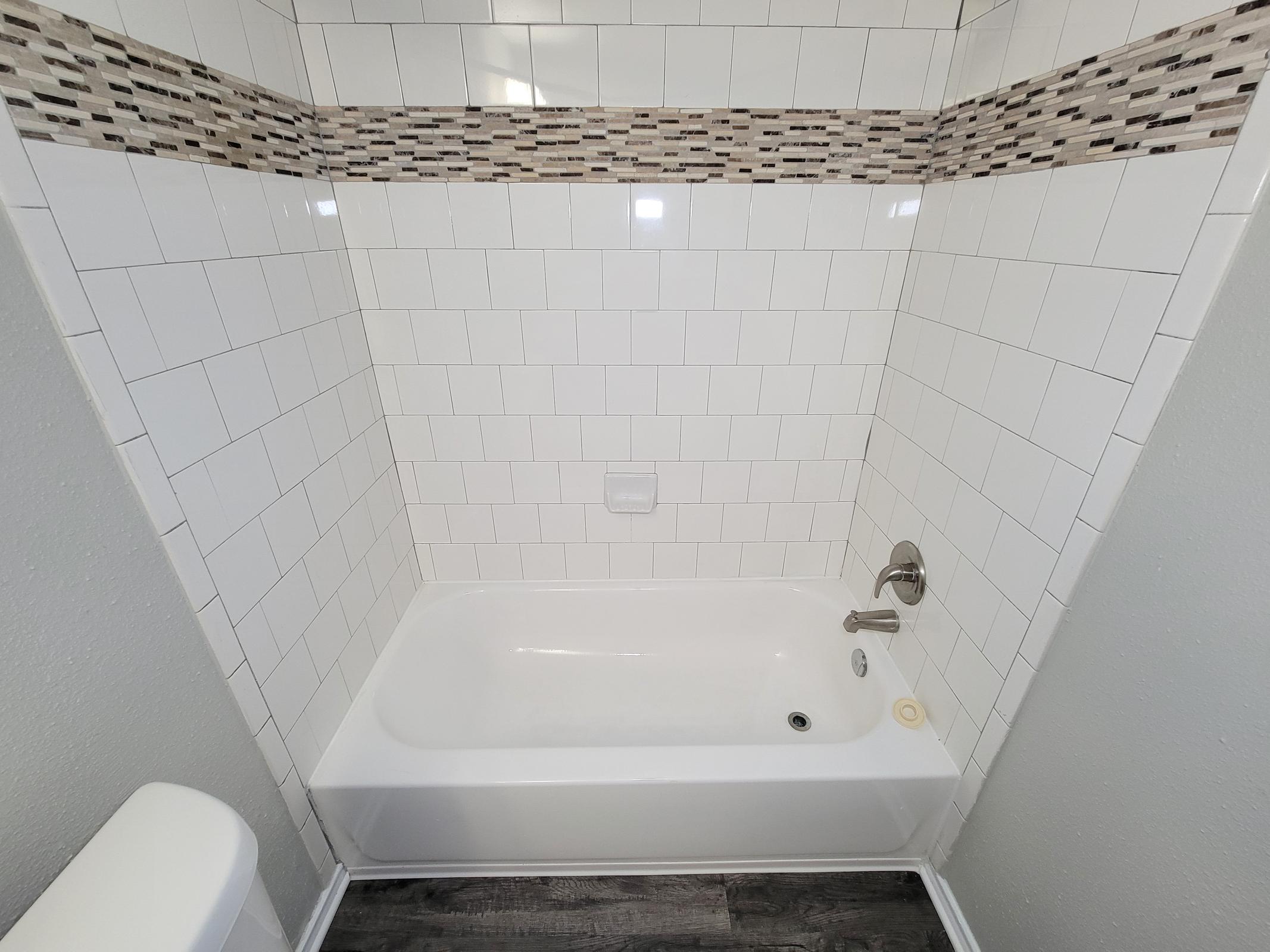 A clean, modern bathroom featuring a white bathtub surrounded by white and patterned tiled walls. The bathtub is positioned against a wall with a decorative tile border. The floor has a dark wood-like finish, and a white toilet is visible to the left.
