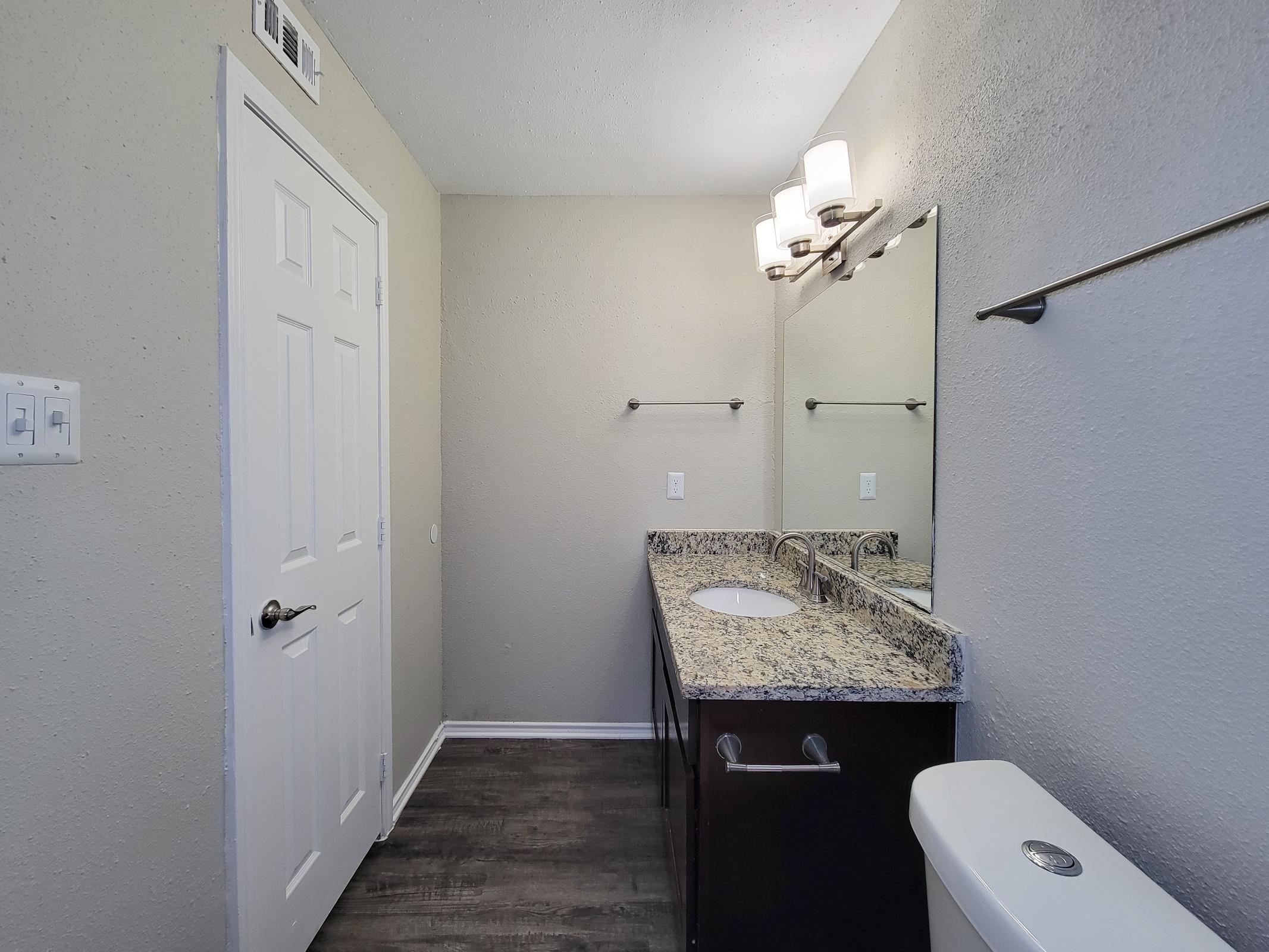 A well-lit bathroom featuring a double vanity with a granite countertop, a large mirror above, and modern light fixtures. There's a white toilet and a door leading to another room, with wood-like flooring and neutral-colored walls.