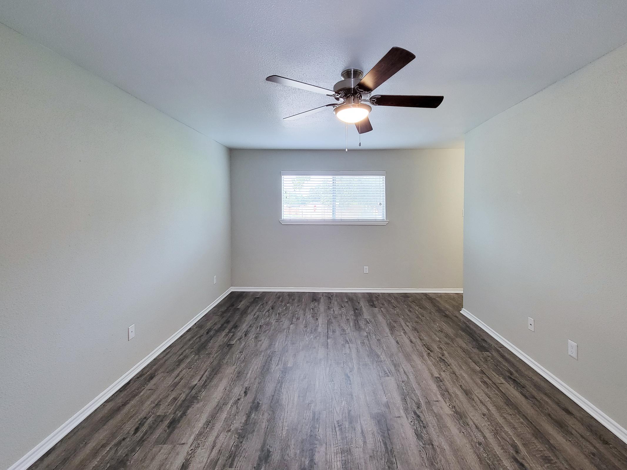 A spacious, empty room with light gray walls and a modern ceiling fan. Natural light comes through a window with blinds. The flooring is a dark, wood-like laminate, creating a clean and fresh atmosphere. The design is simple and minimalist, ideal for various purposes.