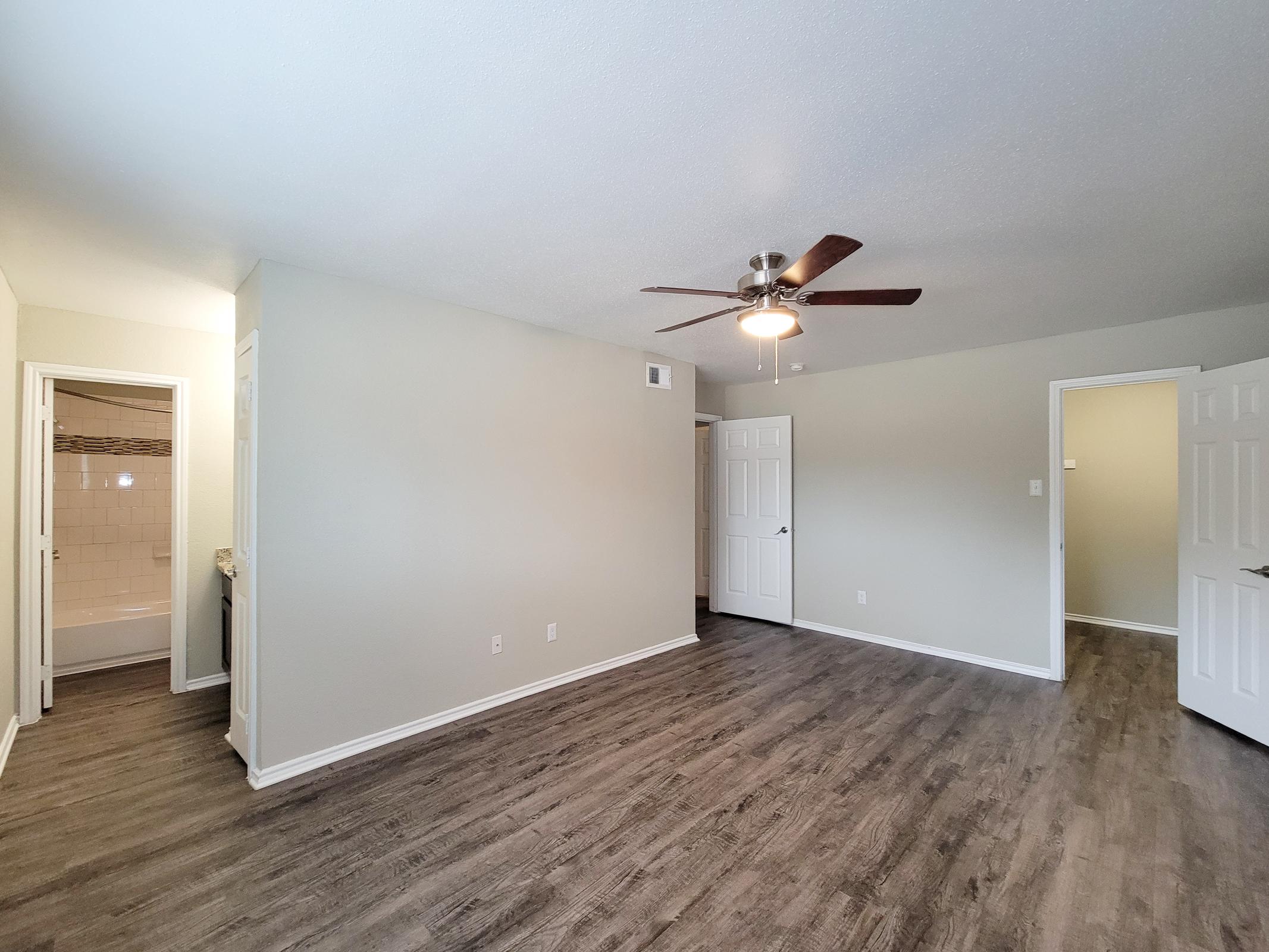 Spacious interior of a room featuring light-colored walls, a ceiling fan with five blades, and modern vinyl flooring. Two doorways lead to adjacent rooms, one of which has a bathroom. Natural light brightens the space, creating an inviting atmosphere.