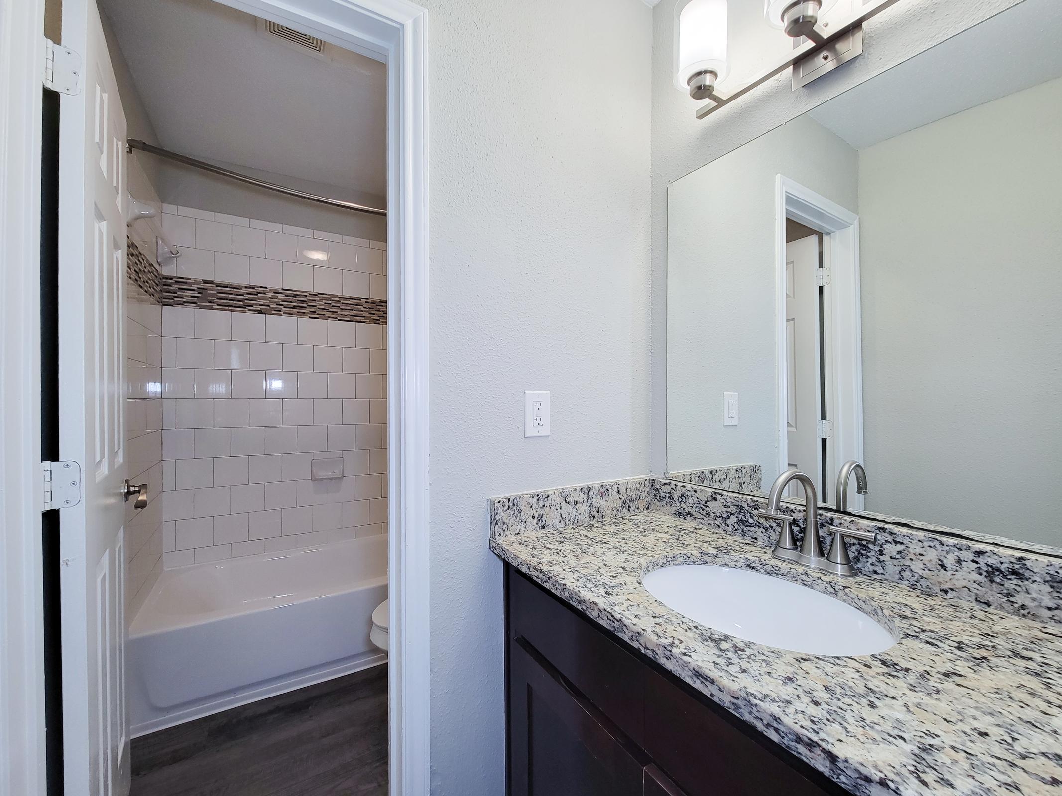 A modern bathroom featuring a double door leading to a shower area with white tiles and a bathtub. The vanity has a granite countertop with a sink and chrome faucet. A large mirror is mounted above the sink, and there is soft lighting from wall-mounted fixtures. The walls are painted in a light color, enhancing the space's brightness.