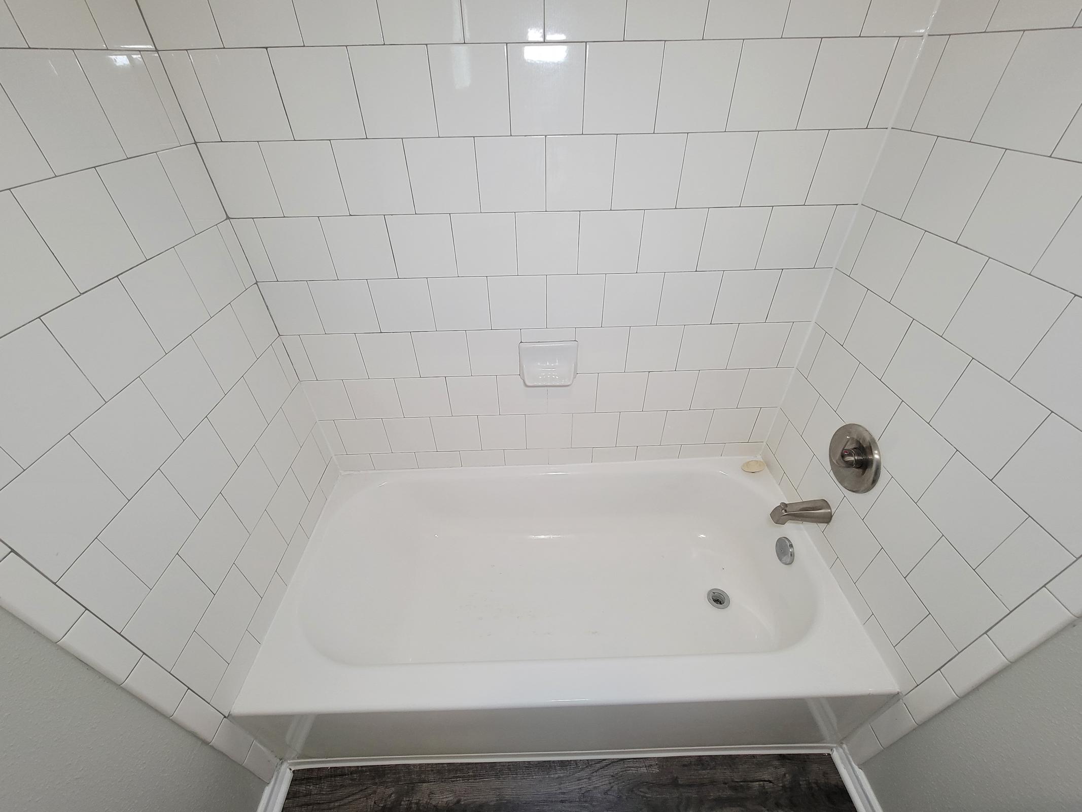 A clean, white tiled bathroom featuring a rectangular bathtub with a chrome faucet and a soap dish on the wall. The flooring is dark, contrasting with the bright, white tiles. The overall look is modern and minimalistic.