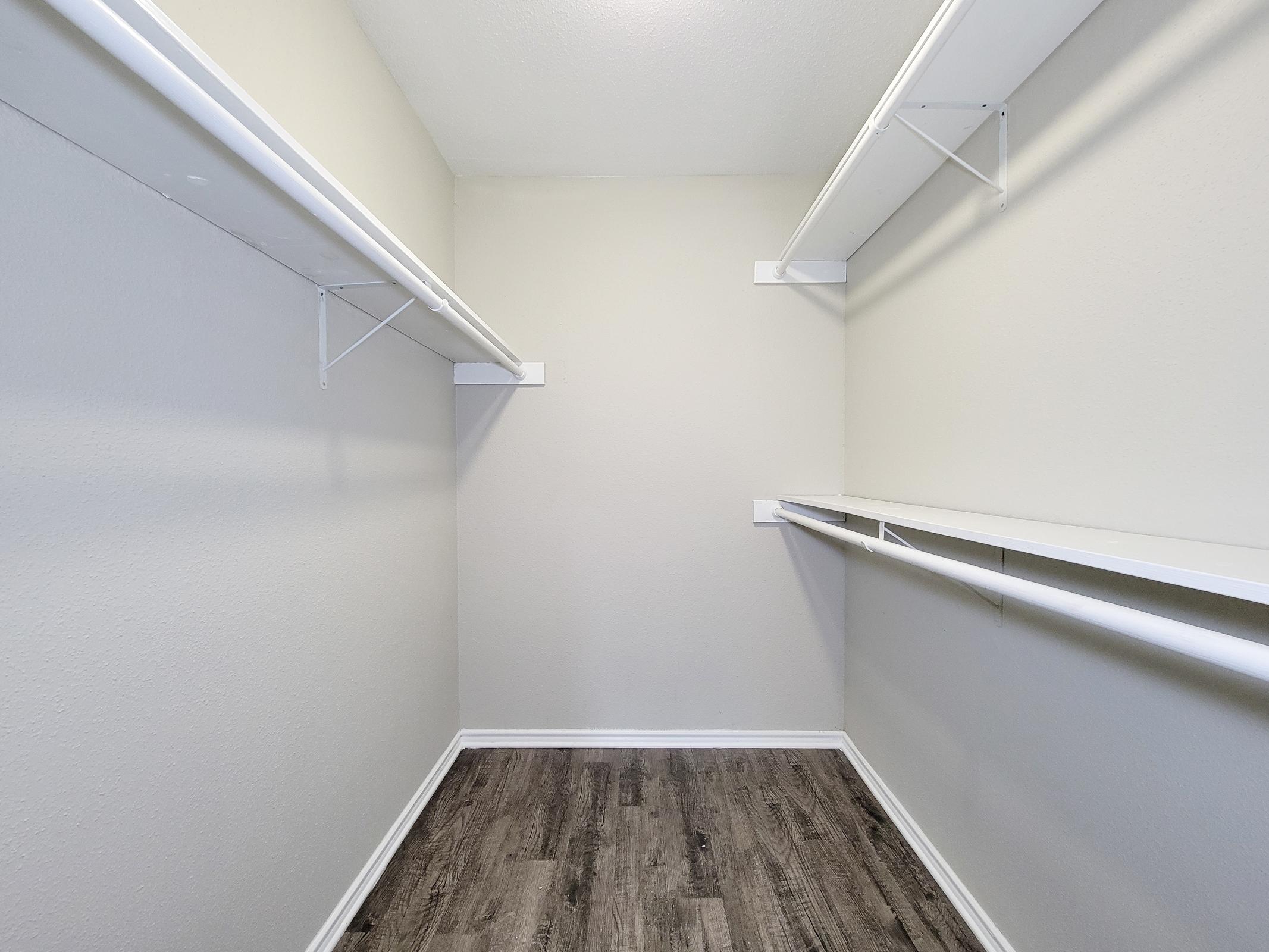 A clean, empty closet featuring light gray walls, a wooden floor, and white shelving. The space has no items inside and is well-lit, providing a minimalist appearance that highlights its spaciousness.