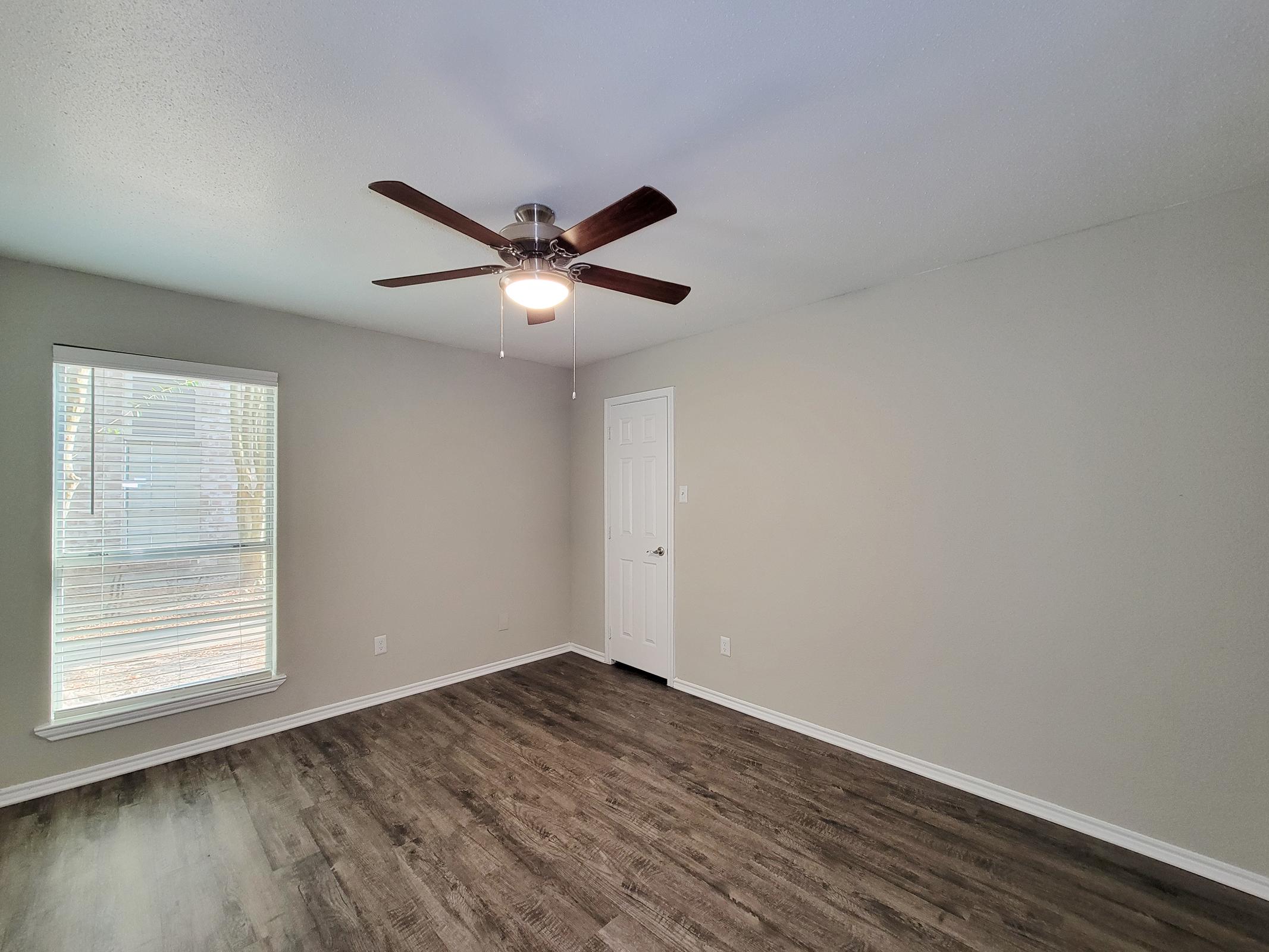 A well-lit interior space featuring a ceiling fan, light gray walls, and laminate flooring. There is a window with blinds allowing natural light, and a closed door. The room appears empty and spacious, suitable for various uses such as a living room or bedroom.