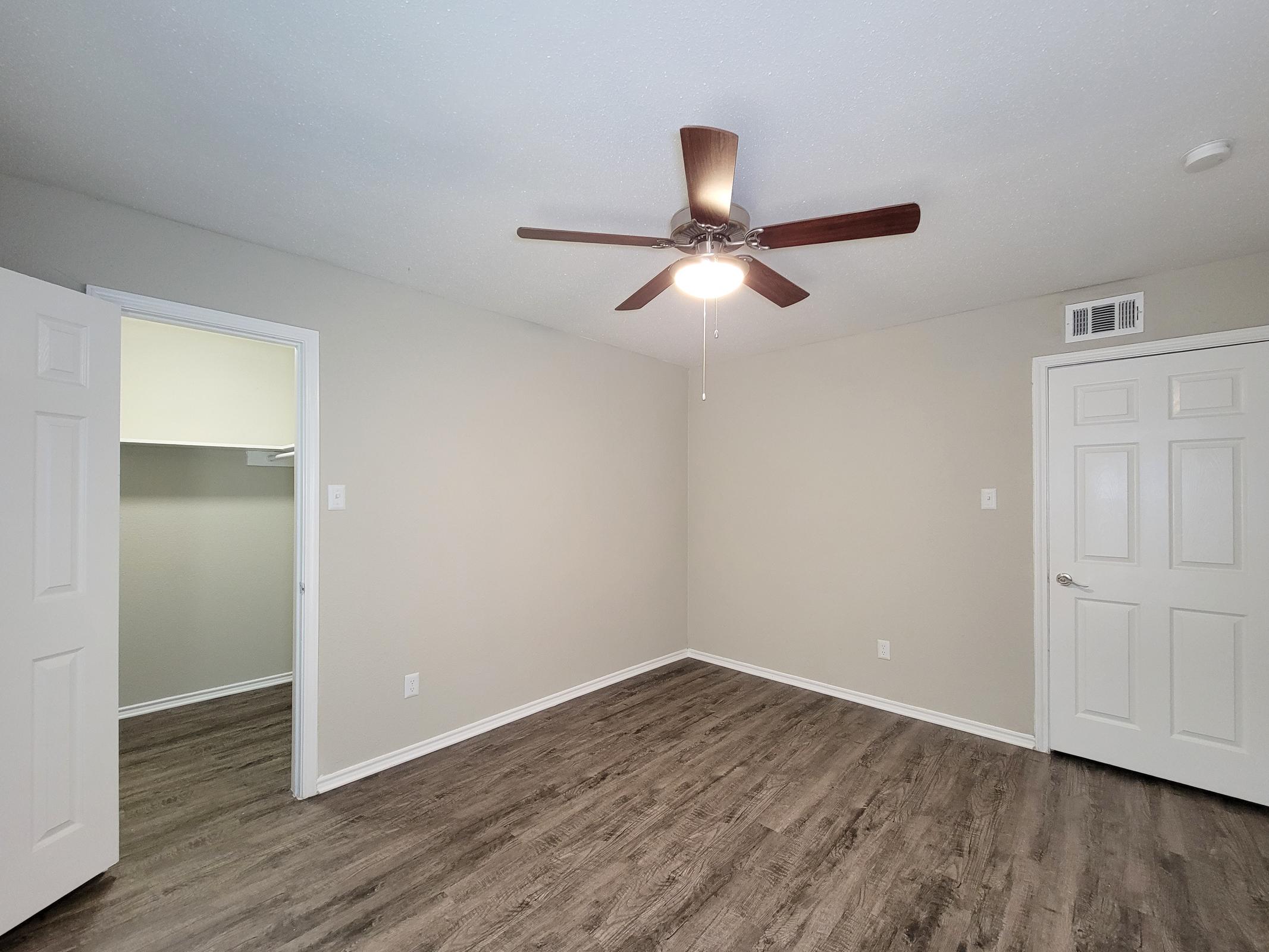 A spacious, well-lit room featuring a ceiling fan, light-colored walls, and dark wood-like flooring. There are two white doors, one leading to a closet with ample space and the other as an entrance/exit. The overall ambiance is modern and inviting.