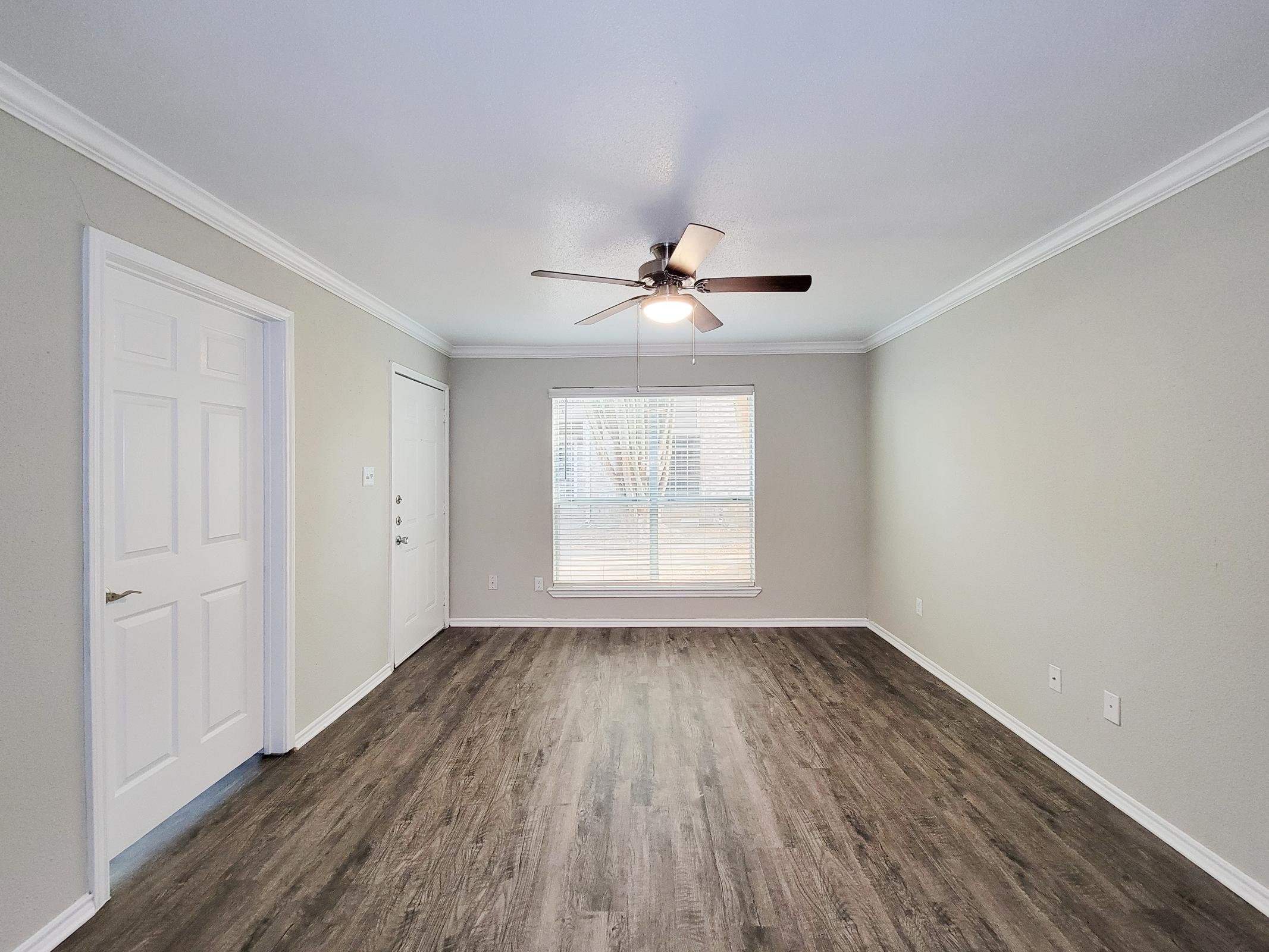 Spacious living room featuring a ceiling fan, light-colored walls, and large window with blinds allowing natural light. The room has stylish wooden flooring and is adjacent to a door leading outside, with a closet on the left side.