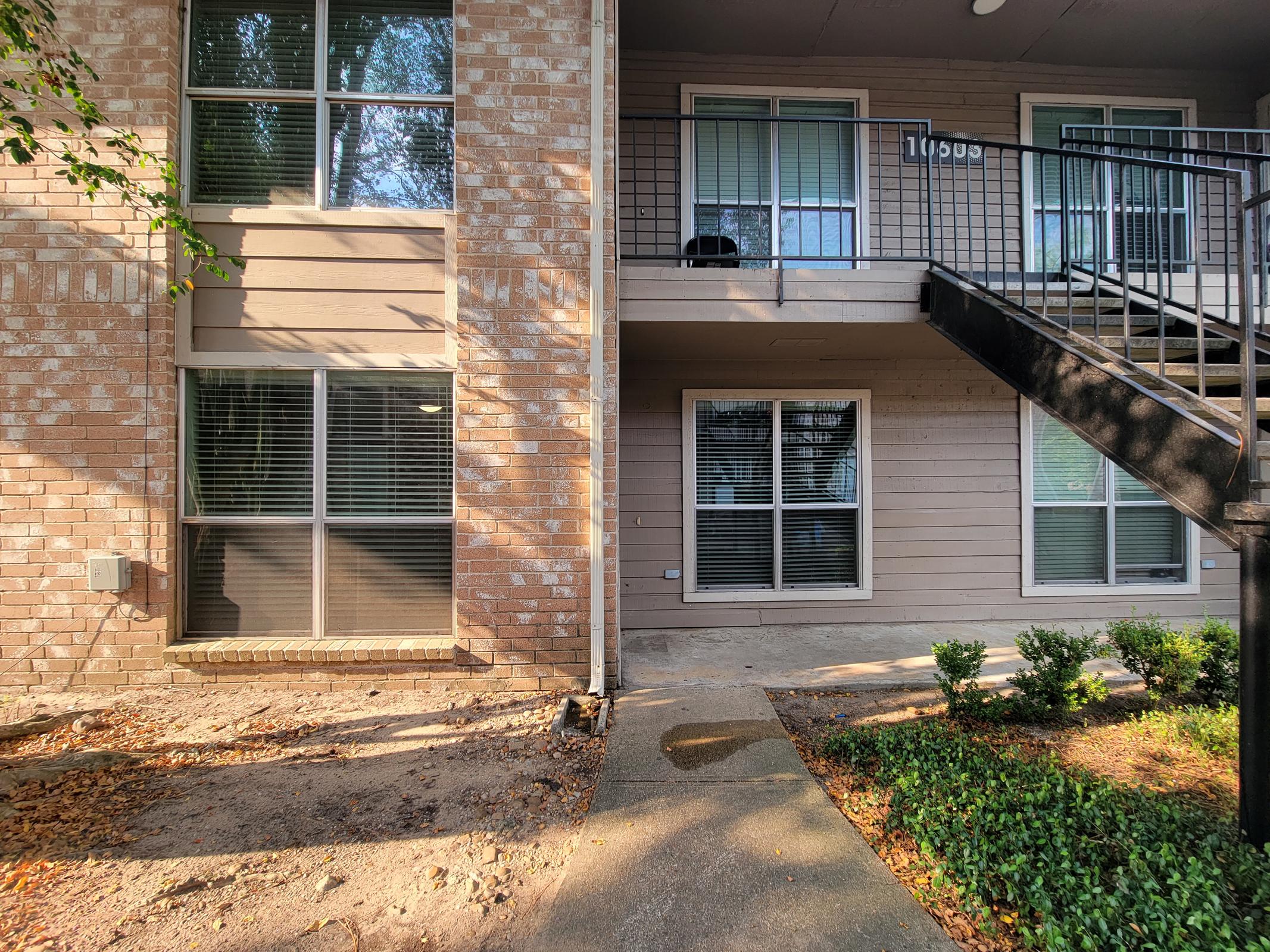 View of a two-story apartment building entrance, featuring beige brick and siding. There are large windows with blinds and a black railing staircase. The ground is a mix of concrete and dirt, with small bushes along the walkway. Sunlight casts shadows on the façade, creating a bright outdoor scene.
