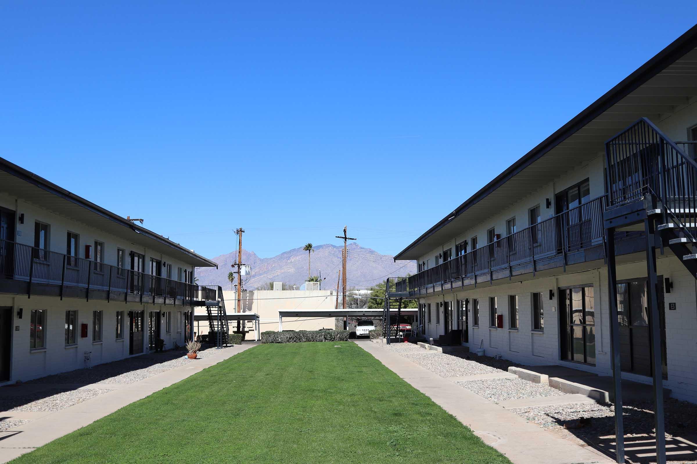 A view of a two-story motel featuring a grassy courtyard in the center. There are walkways leading up to rooms on either side, with a clear blue sky above and distant mountains in the background. The scene is well-lit and conveys a peaceful atmosphere.