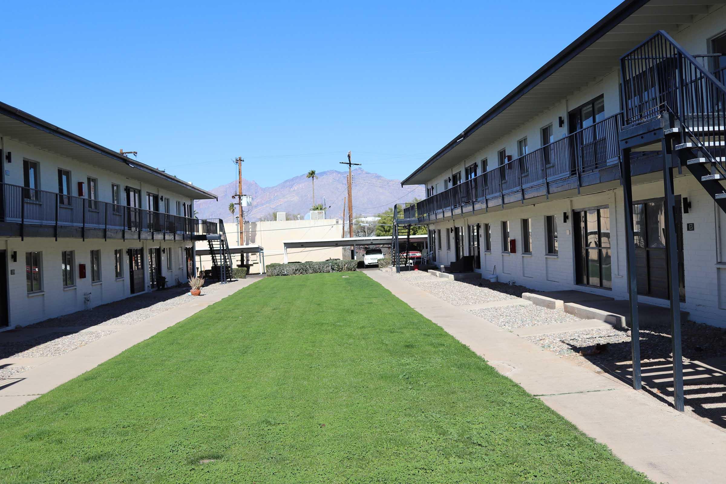 A view of a two-story motel complex with a grassy courtyard in the middle. The buildings have balconies and are arranged in a U-shape, with a clear blue sky and distant mountains in the background. The pathway is lined with gravel, and there are stairs leading to the upper floors.