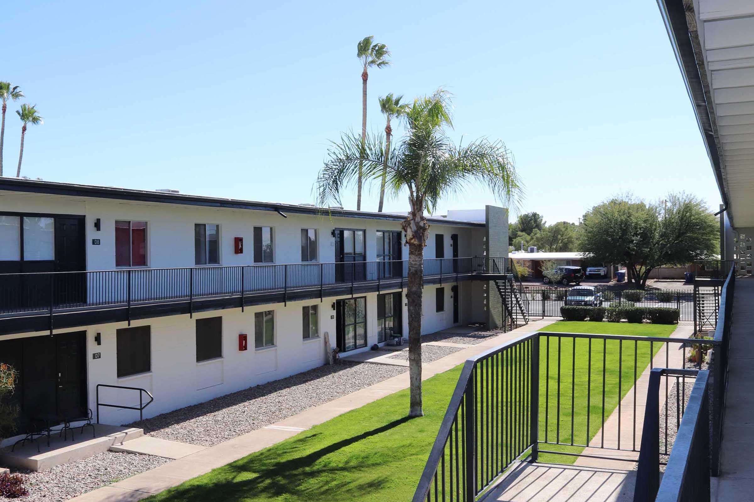 View of a multi-story apartment complex featuring a landscaped courtyard with grass and palm trees. The building has multiple balconies and a staircase visible. The scene is bright and sunny, showcasing the exterior of the apartments and the surrounding area.