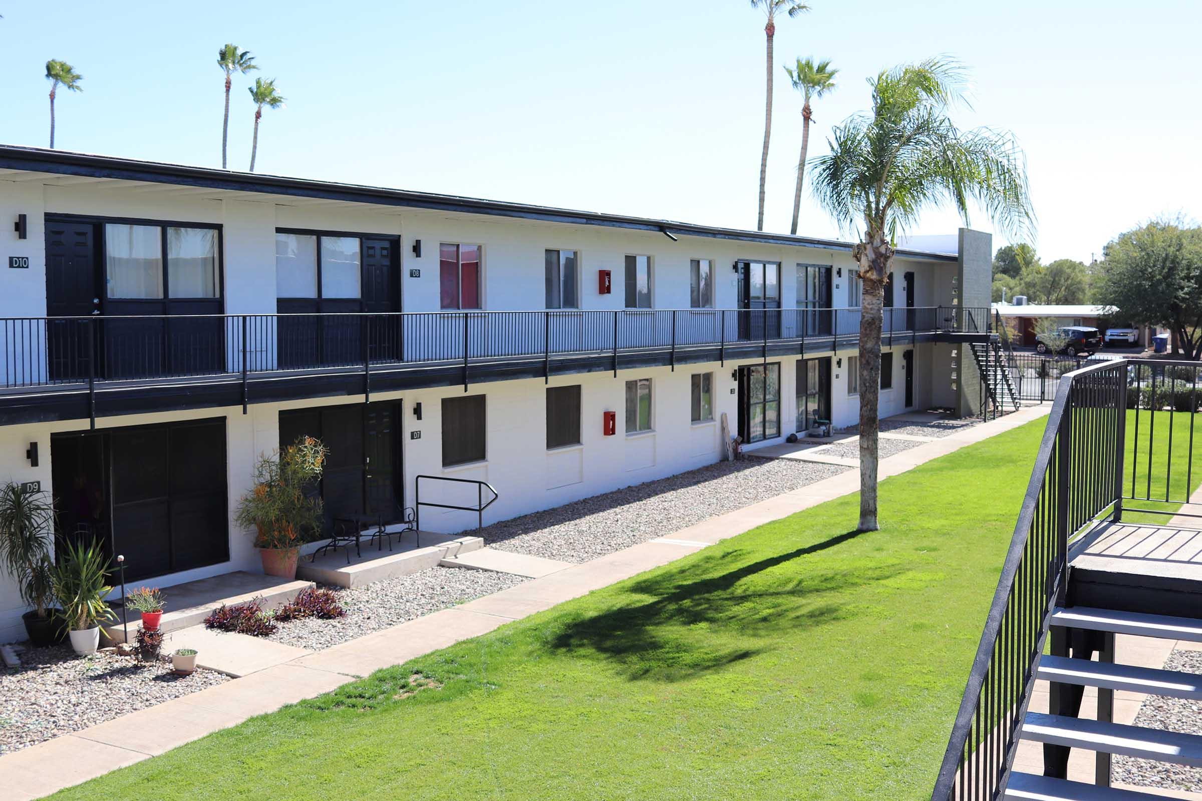 Two-story apartment building with multiple units, featuring a balcony on the upper level. The pathway is lined with decorative stones and a small garden area, with palm trees and green grass in the foreground. Clear blue sky above, indicating a sunny day.