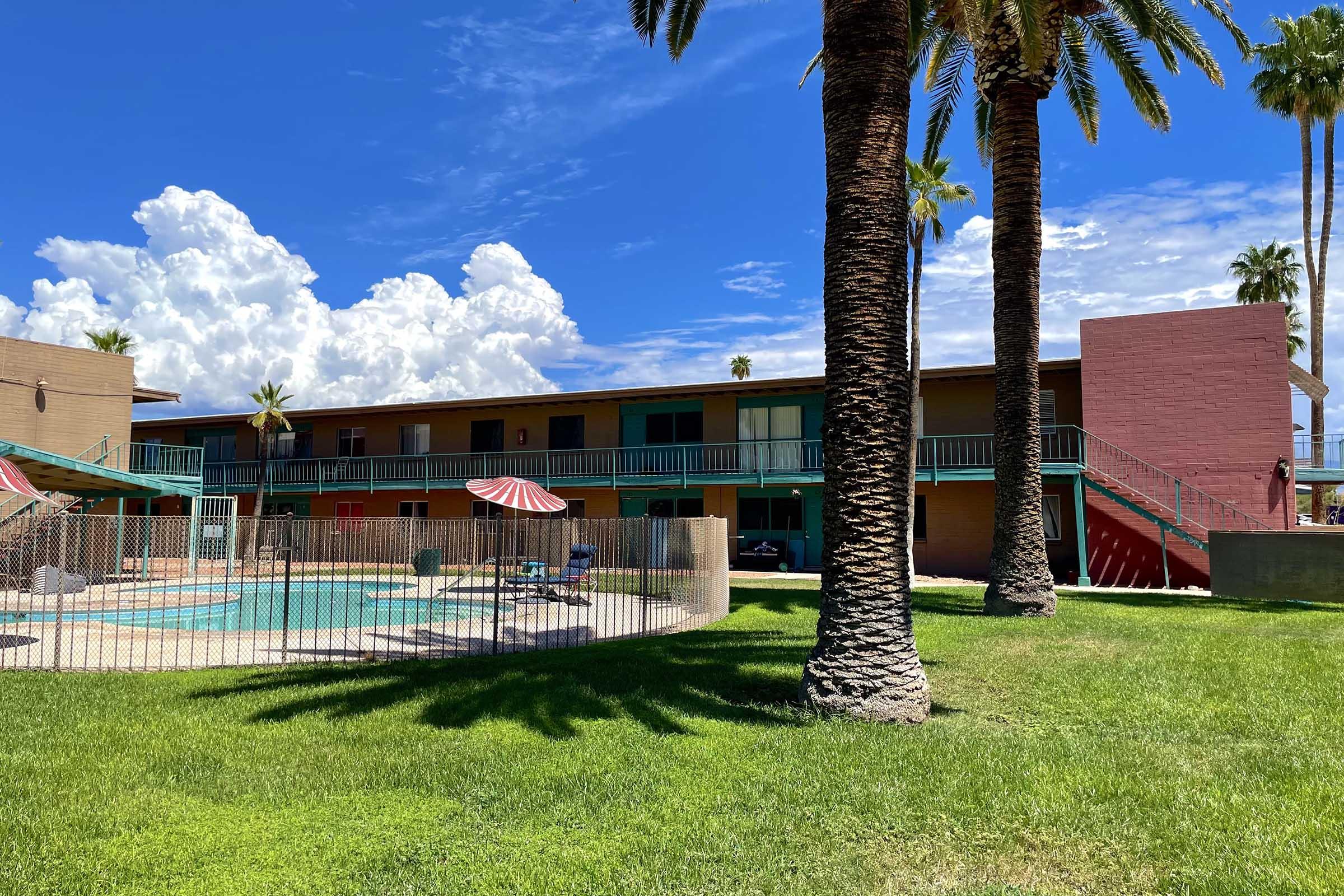A sunny view of a palm tree-lined courtyard featuring a swimming pool surrounded by a fence. In the background, a two-story apartment building with green balconies and a reddish wall is visible against a blue sky filled with fluffy white clouds.