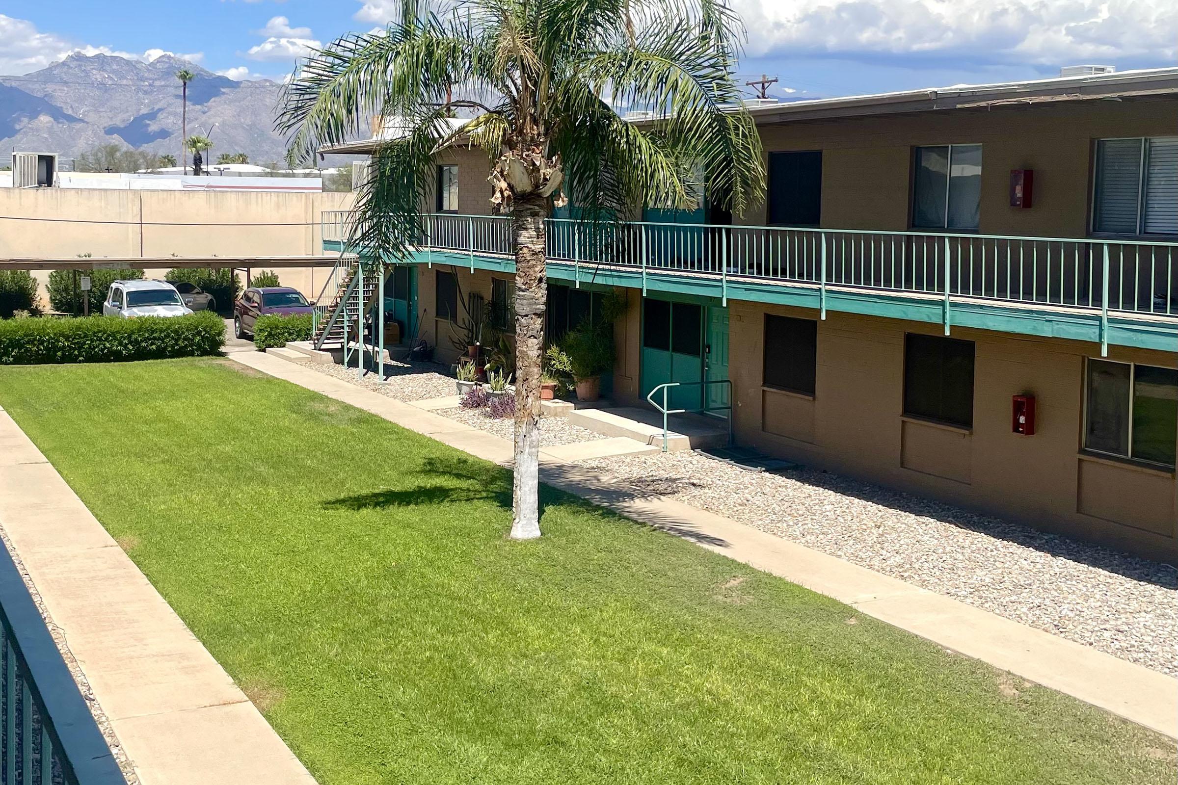 A view of a courtyard at an apartment complex featuring a well-maintained lawn, a palm tree, and two levels of balconies. The background shows mountains under a partly cloudy sky. There are walkways and some outdoor furniture visible, creating a relaxing residential atmosphere.