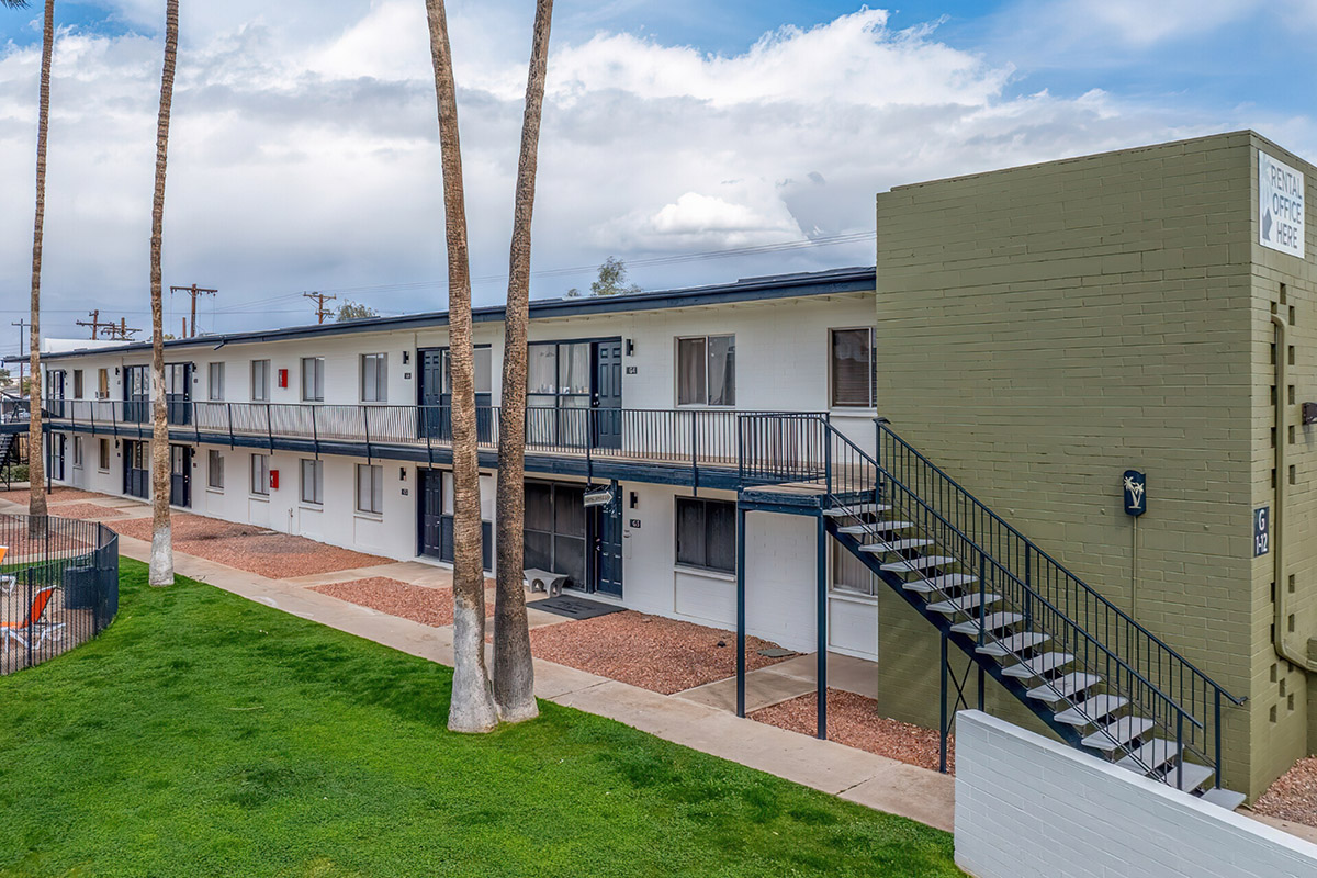 A two-story apartment complex with a green accent wall, featuring multiple units with balconies. There is a staircase leading to the upper level on one side. The grounds are landscaped with grass and gravel, and palm trees are visible in the foreground against a cloudy sky.