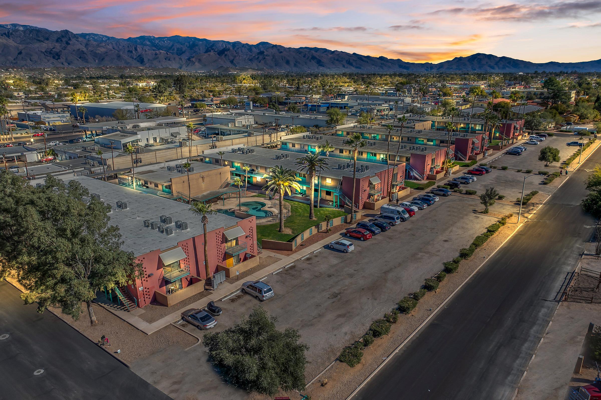 Aerial view of a residential area featuring colorful low-rise buildings with palm trees and a swimming pool. The scene is set during twilight, showcasing a parking lot filled with cars and a backdrop of mountains in the distance under a gradient sky. The ambiance is calm and inviting.