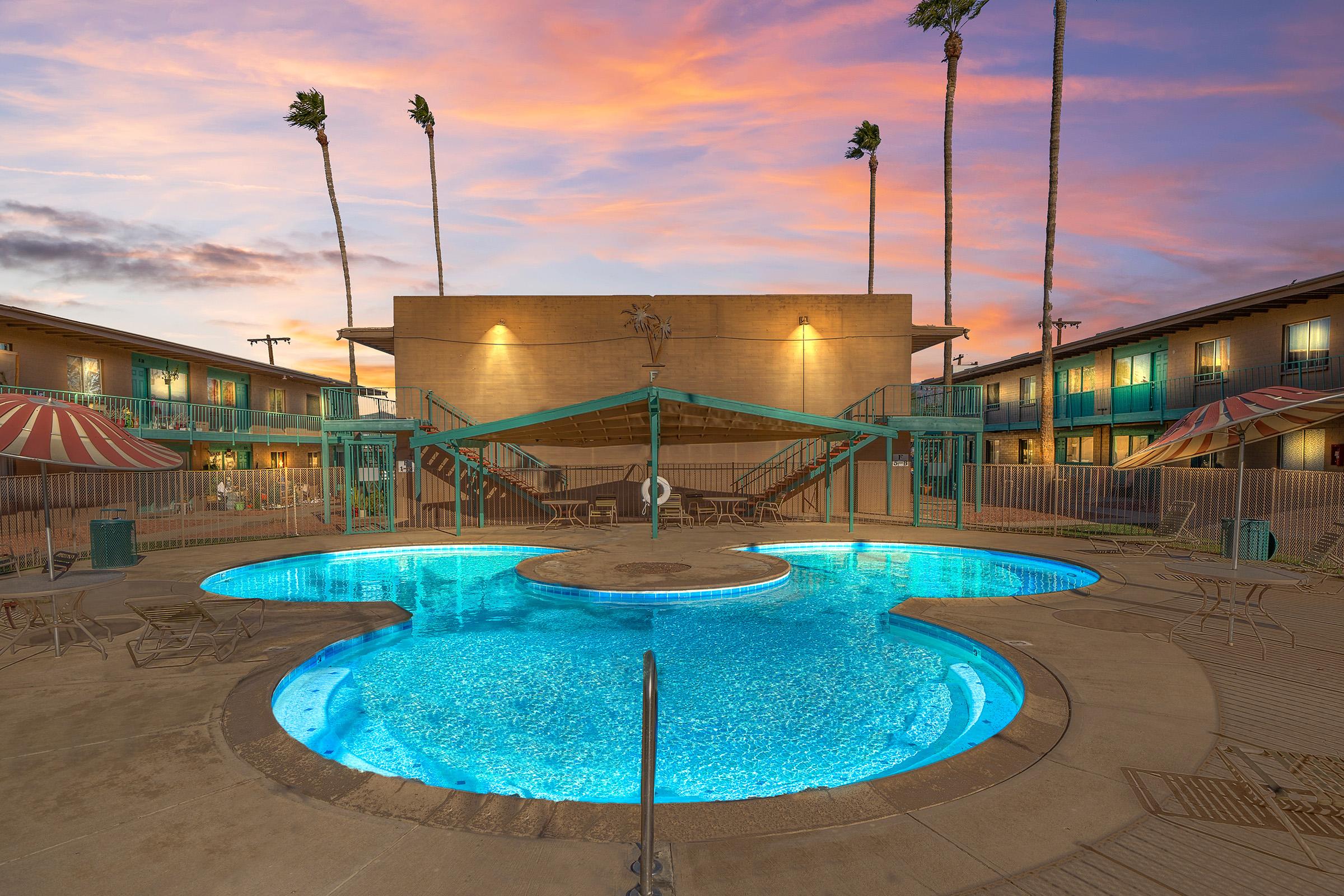 A pool area with two distinct sections, illuminated at dusk. Surrounding the pool are lounge chairs and umbrellas, while palm trees rise in the background against a colorful sunset sky. A two-story building is visible, with balconies overlooking the pool.