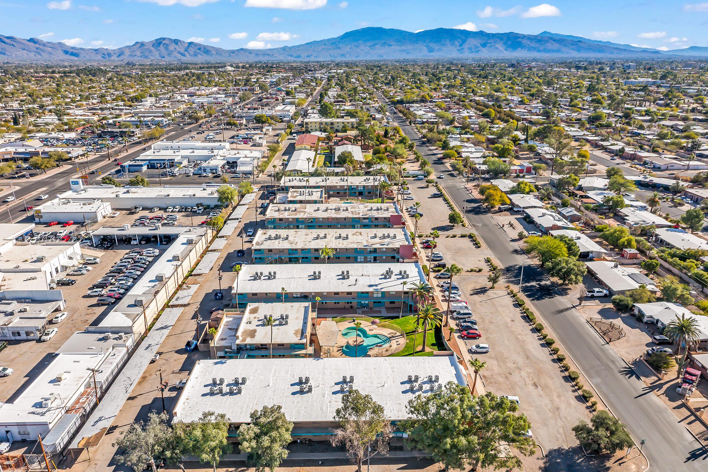 Aerial view of a suburban area featuring residential buildings, a swimming pool, and a street lined with palm trees. Surrounding mountains are visible in the background. The scene includes parking lots and commercial structures, showcasing a mix of urban development and open spaces.