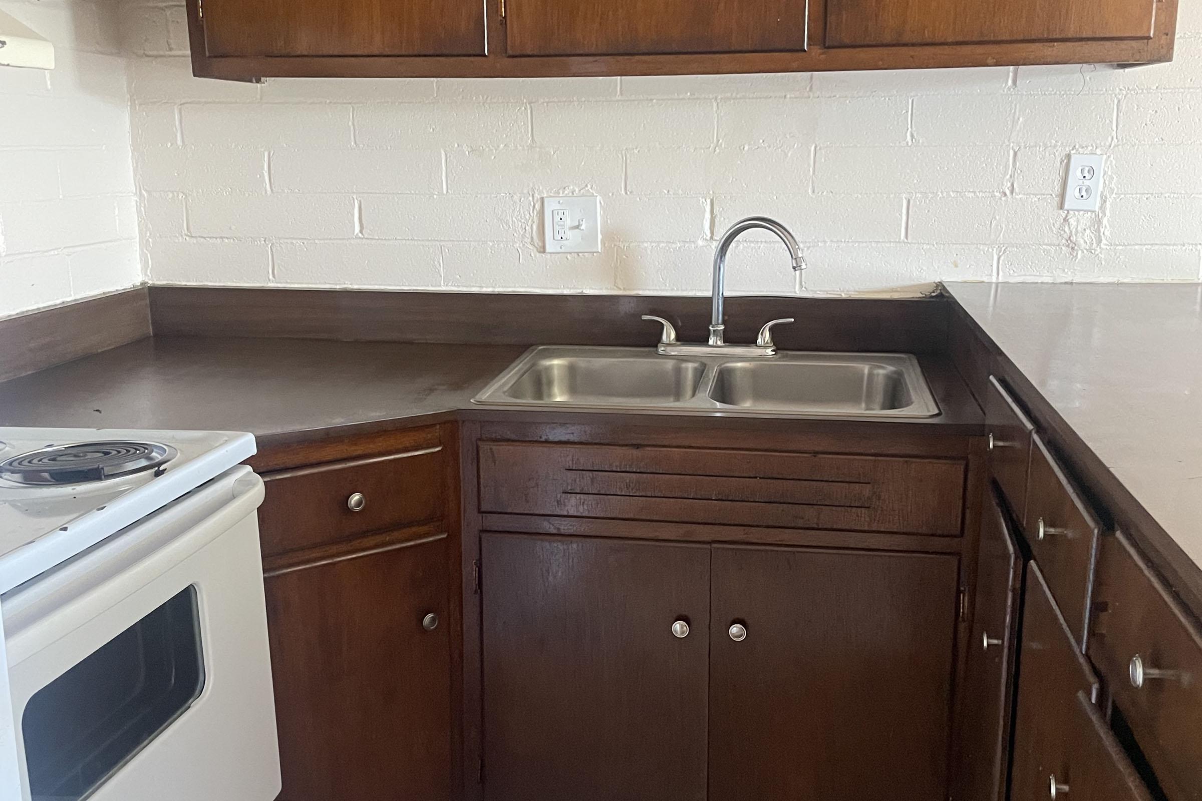 A corner kitchen with dark wooden cabinets and a stainless steel double sink. A white stove is visible on the left side, with a countertop made of dark material. The walls are painted white, and there are electrical outlets nearby. The overall setting is clean and simple.
