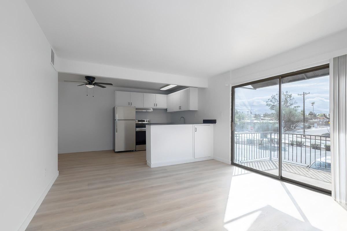 Modern open-concept apartment interior featuring a kitchen with stainless steel appliances, a ceiling fan, and large windows allowing natural light. Light-colored flooring complements the minimalist design, with a balcony view visible through sliding glass doors.