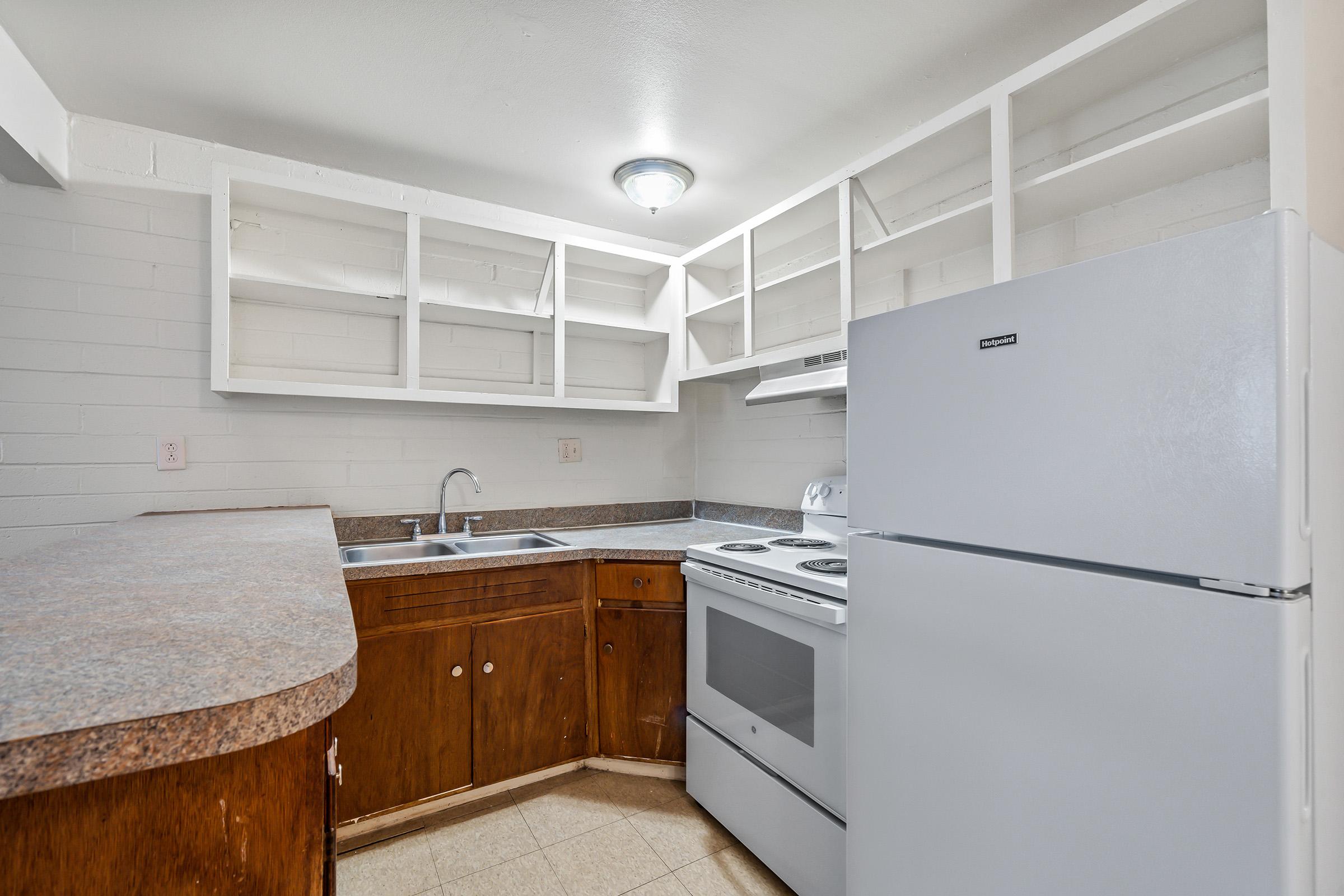 A small kitchen featuring light-colored walls and wooden cabinetry. There is a white refrigerator and an electric range with an oven. The countertop is made of a brown material, and there are open shelves above the sink, providing storage space. The flooring is light-colored tile.