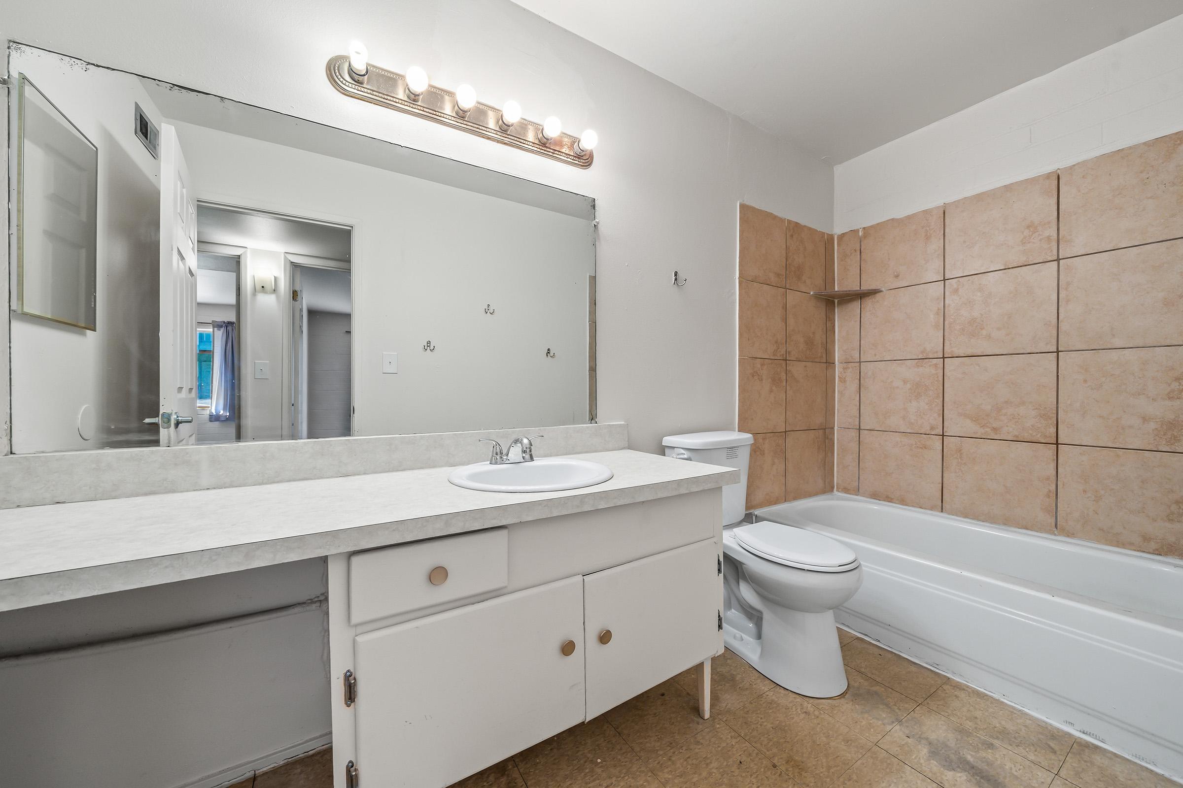 A simple bathroom featuring a bathtub, toilet, and a vanity with a sink. The walls are mostly tiled with light-colored tiles, and there is a mirror above the sink with a light fixture. The floor is light-colored, and the overall decor is minimalistic.