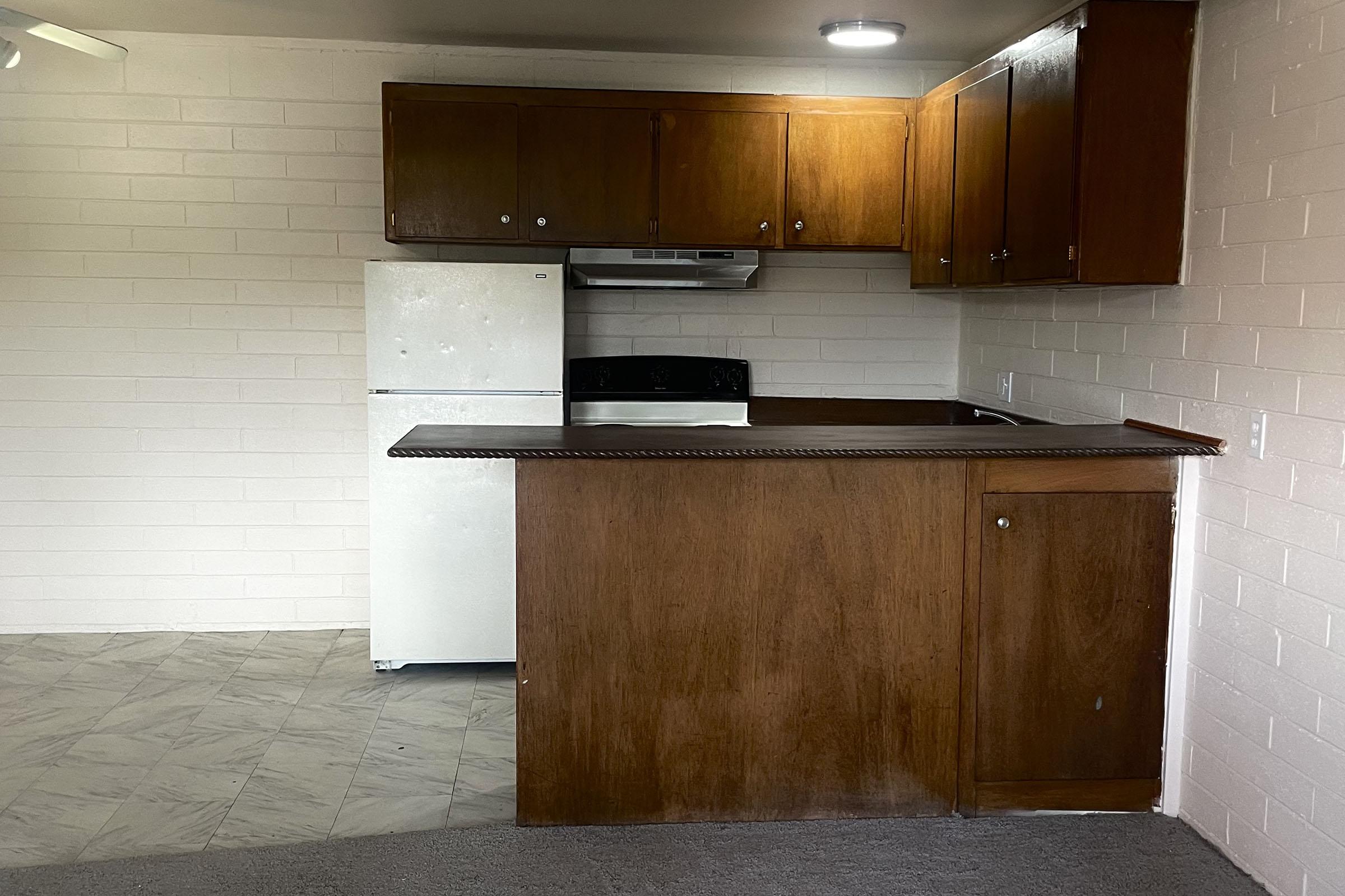 A small kitchen area featuring a wooden countertop with a built-in cabinet, a white refrigerator, and a black stove with a hood. The walls are painted white brick, and the flooring is a mix of tile and carpet. There are wooden cabinets above the countertop providing storage space.