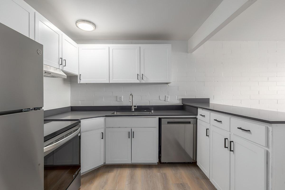Modern kitchen featuring white cabinets, a stainless steel refrigerator, a black countertop, and a built-in sink. The layout includes a stove and a dishwasher, with light-colored walls and wooden flooring enhancing the bright, clean aesthetic.