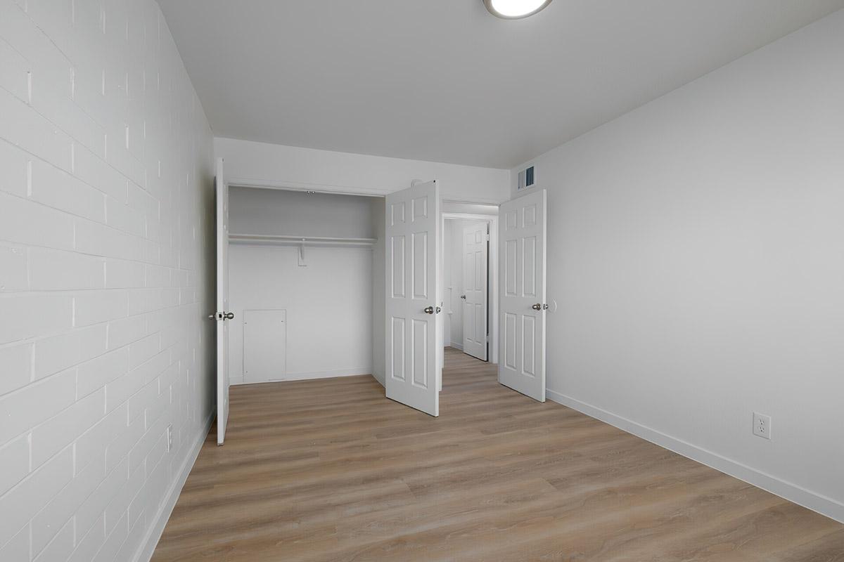 Empty room featuring light-colored walls and wooden flooring. Two open doors lead to a closet and another room, with natural light coming through. The interior appears modern and minimalistic, suitable for various uses or decoration.