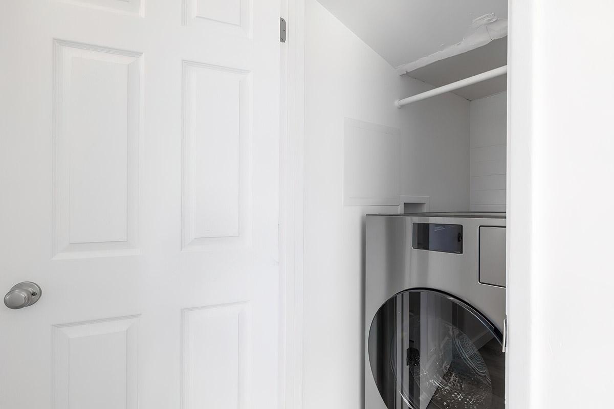A modern laundry room featuring a stainless steel washing machine and dryer. The room has white walls and a door leading to the hallway, with a simple and clean design. Natural light may come from a nearby window, highlighting the contemporary appliances.
