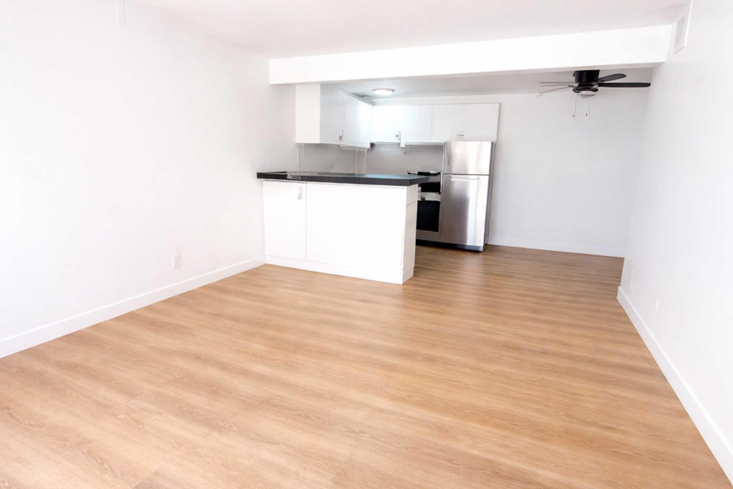 A bright, modern kitchen and living area with light wooden flooring. The kitchen features white cabinetry, a black countertop, and stainless steel appliances. There is a ceiling fan in the corner of the room, and the walls are painted white, creating an open and airy atmosphere.