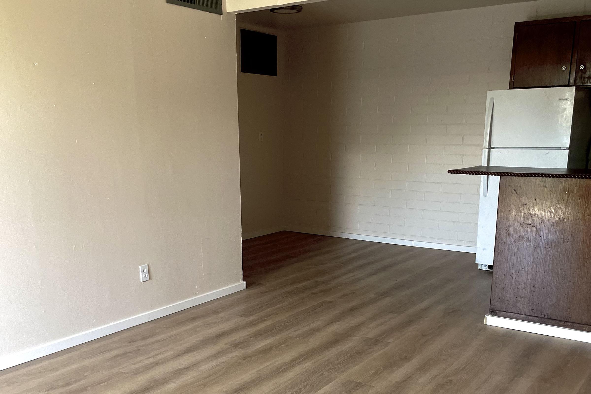 An empty room featuring light-colored walls and a wooden floor. There is a white refrigerator and a wooden counter visible in the kitchen area, with an open space leading to the rest of the room. Natural light comes through a small window.