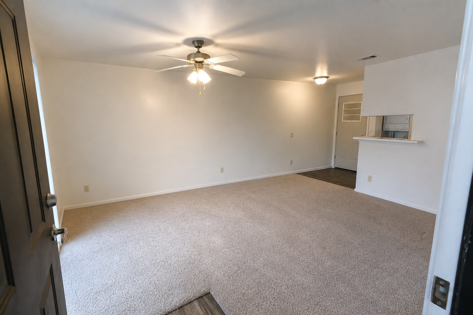A spacious, empty room featuring light beige walls and carpet. A ceiling fan is mounted in the center, and there is a small counter opening leading to another area. The door is visible in the foreground, and the light fixtures create a warm ambiance.