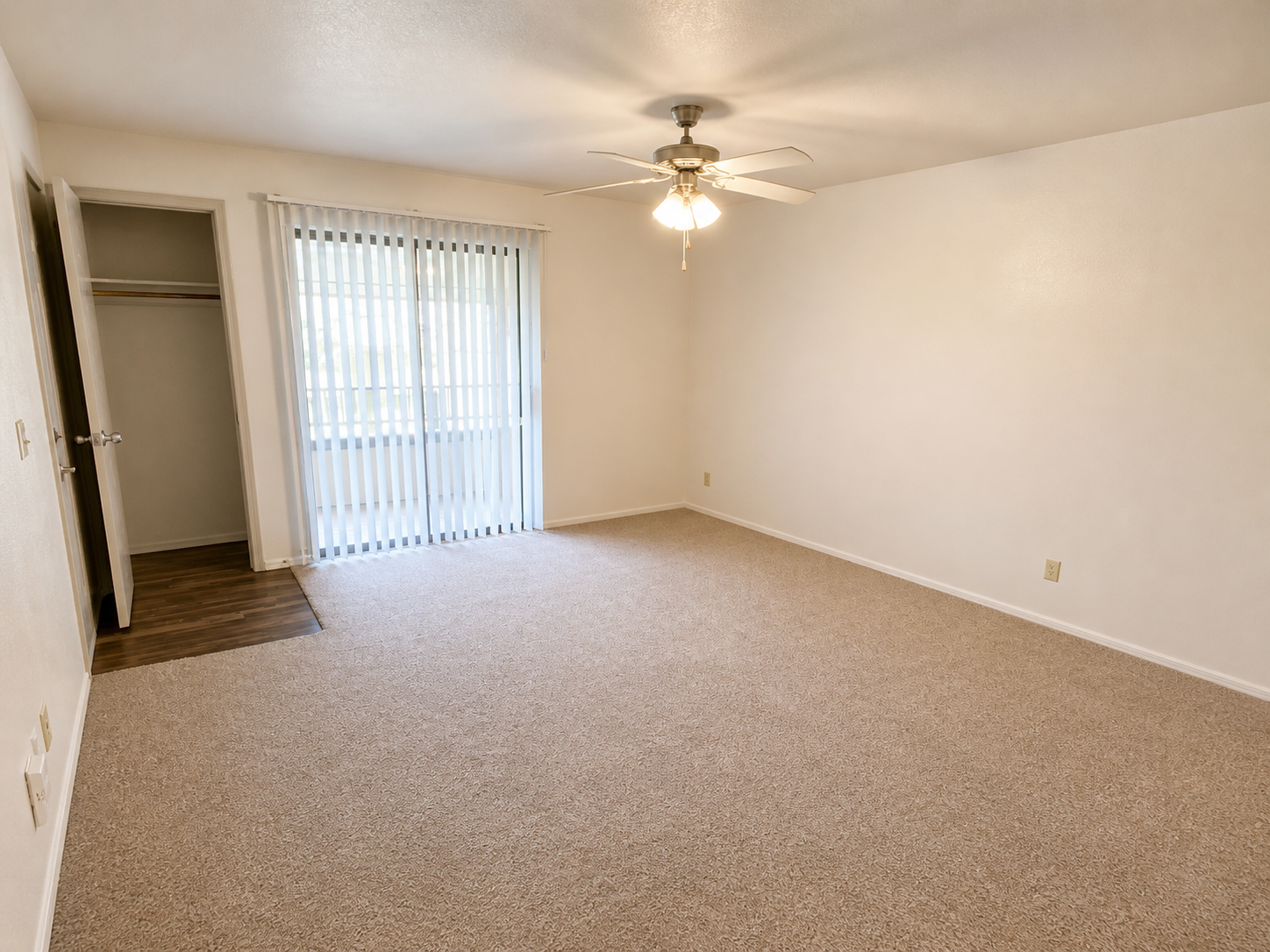 A spacious, empty room with light beige carpet and white walls. It features a ceiling fan and large sliding glass doors leading to a balcony or patio with vertical blinds. There is a closet on the left, and the overall ambiance is bright and inviting.