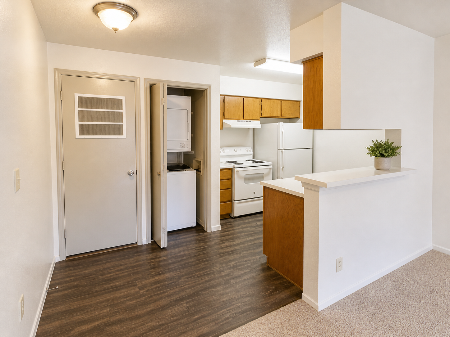 A bright, modern kitchen and living area in an apartment. The kitchen features wood cabinets, a stove, refrigerator, and a laundry area. There's a small bar counter separating the kitchen from the living space, with light-colored walls and a ceiling light. The flooring combines hardwood and carpet for a cozy feel.