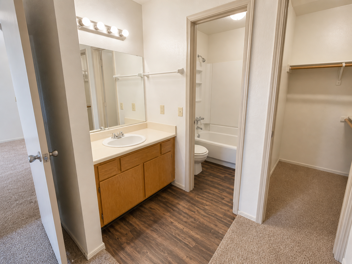 A clean, well-lit bathroom featuring a single sink vanity with wooden cabinets, a mirror above, a shower-tub combo, and a toilet. The flooring is a mix of laminate and carpet, with an open closet space visible. The walls are painted in light colors, enhancing the bright and spacious feel.