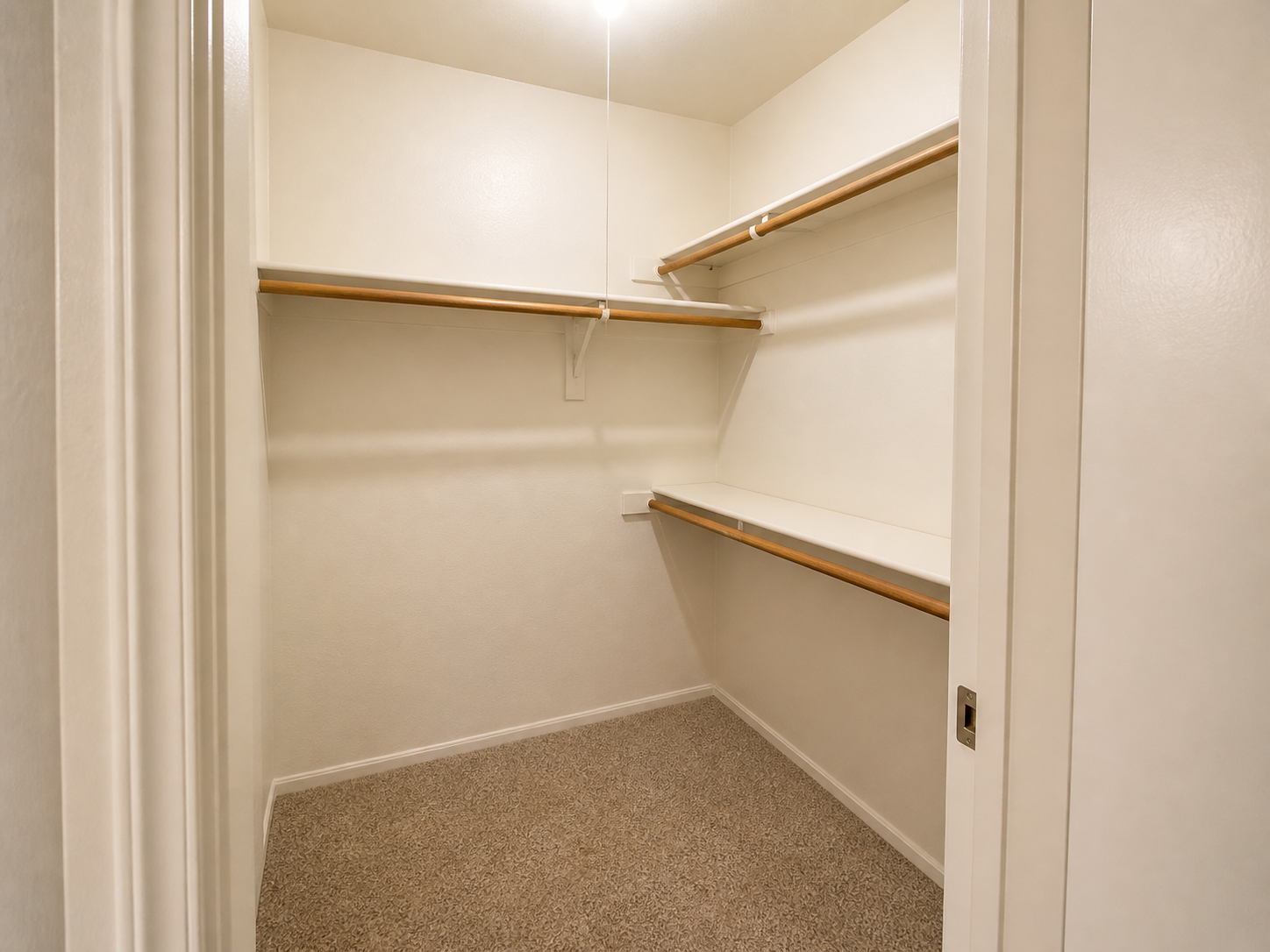 A clean, empty closet featuring two wooden shelves along the walls, with a light fixture hanging from the ceiling. The walls are painted a light color, and the floor is covered with beige carpet. The closet has a simple and spacious layout, ready for storage.