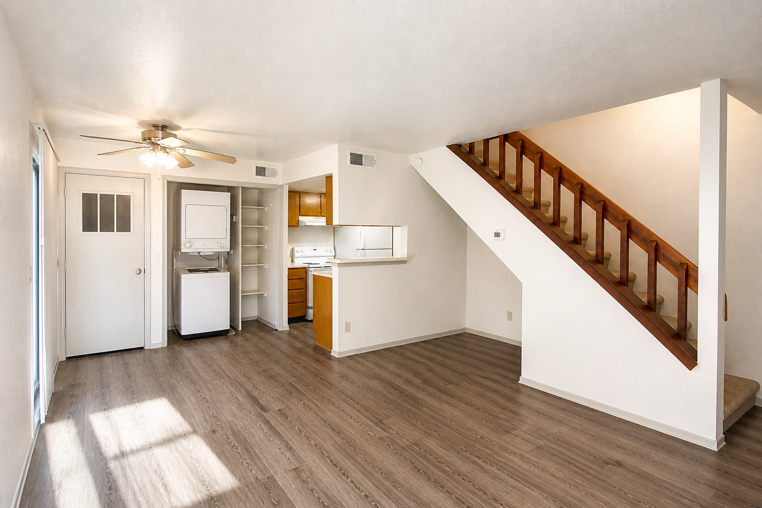 Bright, spacious living area with light-colored walls and wooden flooring. A ceiling fan circulates air, and a staircase leads to an upper level. To the left, a door opens to the outside, while a white washer and dryer are visible near the kitchen area, which features wooden cabinets.