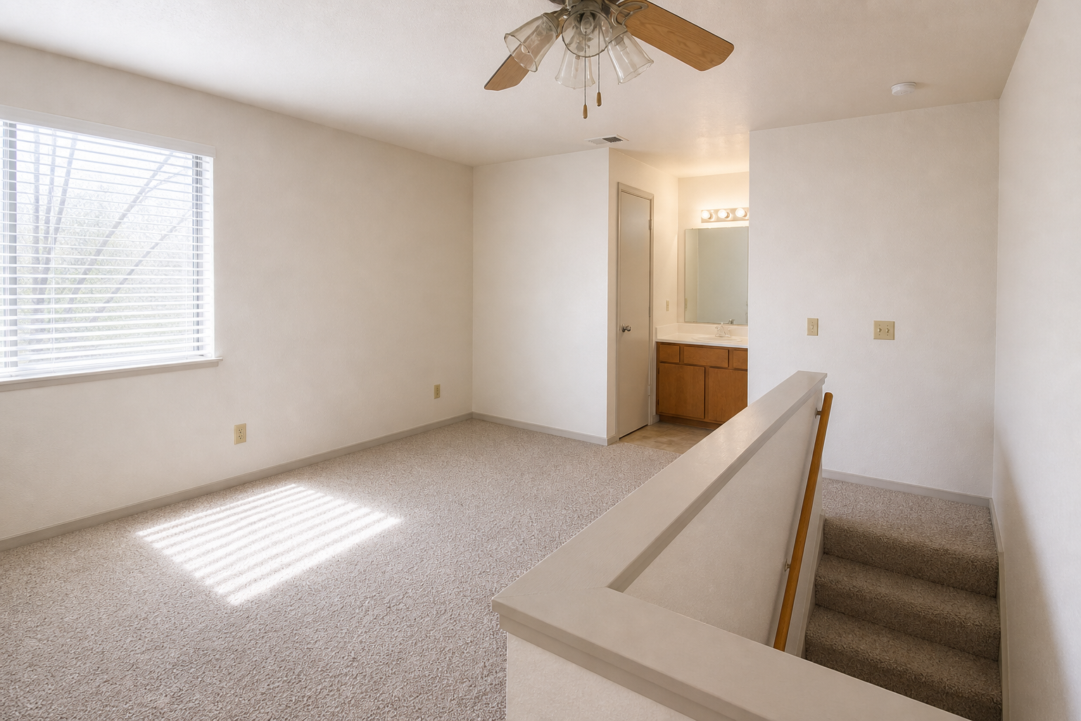 A spacious, light-filled room with beige walls and carpeting. A ceiling fan is mounted above, and a window with blinds allows natural light. To the right, an open doorway leads to a bathroom area with a single vanity and mirror. A staircase is visible in the foreground, leading to another level.