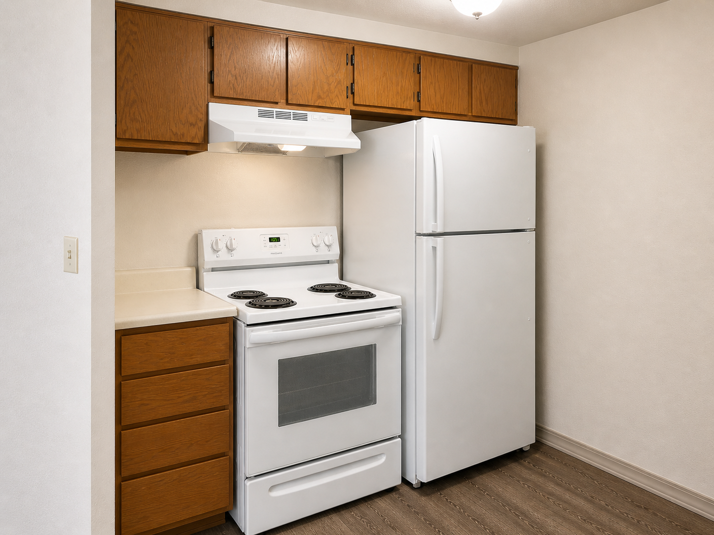 A simple kitchen featuring a white gas stove and oven, a refrigerator, and wooden cabinets. The countertop is plain and light-colored, and the flooring is a light wood laminate. Soft lighting illuminates the space, creating a clean and functional atmosphere.