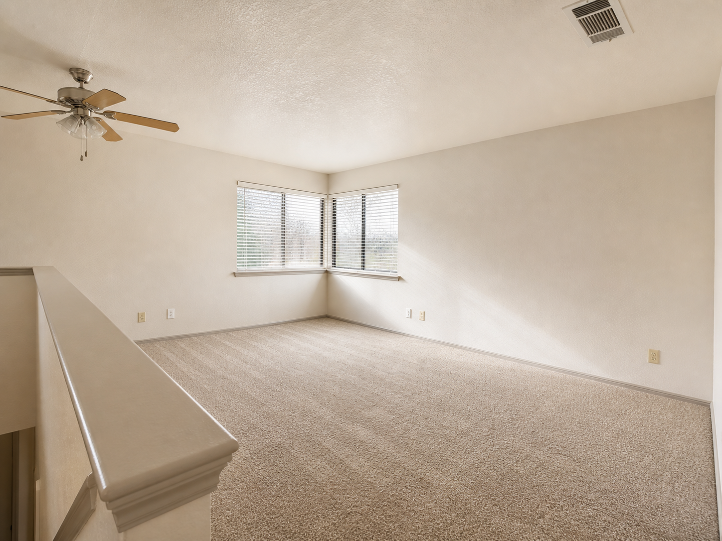 A spacious, empty room featuring light-colored walls and carpet. There is a ceiling fan and a large window with blinds, allowing natural light to enter. The room has a slightly elevated area and is designed to accommodate various furniture arrangements.