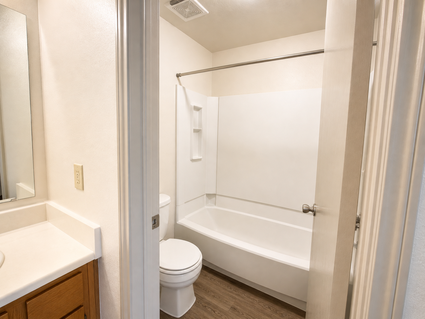 A clean, well-lit bathroom with a bathtub and shower combination against a white wall. It features a white toilet and a vanity with a wooden cabinet. The mirror above the sink reflects the bright ambiance, while the floor is covered in light brown laminate. The doorway frames the view into the room.