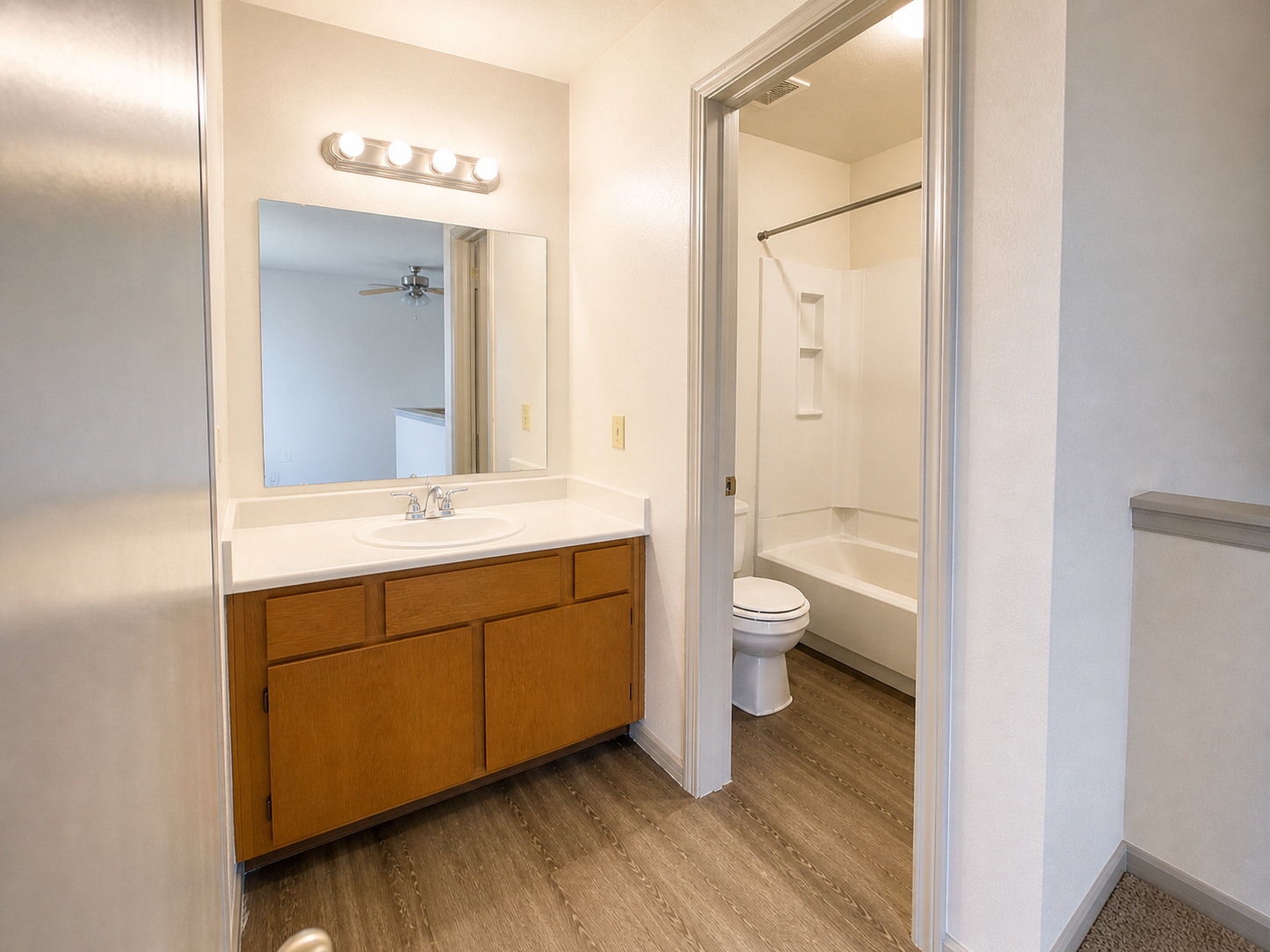 A bright and clean bathroom featuring a wooden vanity with a sink, a large mirror with four light fixtures above, and a framed shower area. The floor is covered in light-colored laminate, and a white toilet is visible in a separate alcove. The walls are painted a soft, neutral color.