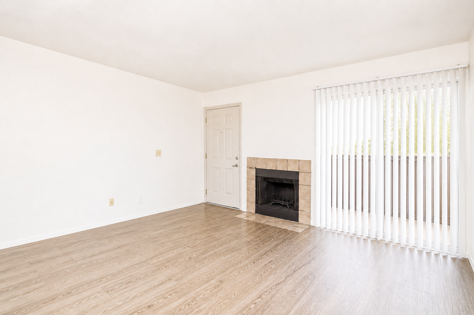 A spacious, bright living room with light-colored walls and wood-look flooring. It features a beige tiled fireplace, a door leading outside, and vertical blinds covering a window. The room is empty, creating a clean and open atmosphere, ideal for personalization.